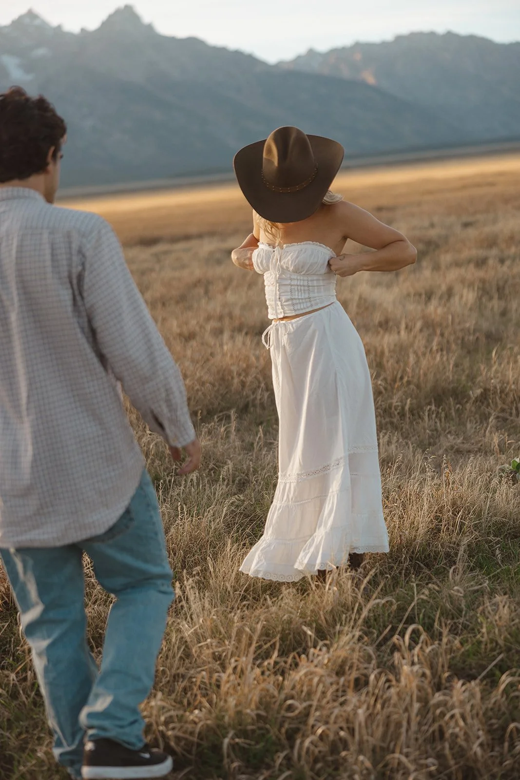 A woman in a white dress and large brown hat adjusting her top in a grassy field with mountains in the background, accompanied by a man in a checkered shirt and jeans.