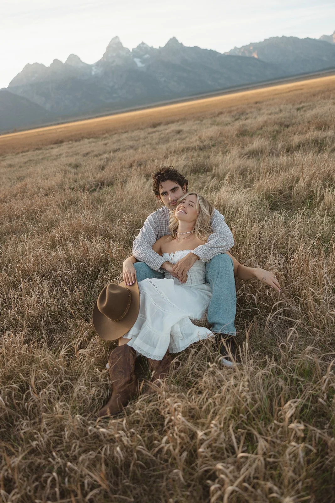 A young couple sitting in a field of tall grass with mountains in the background, the woman is wearing a white dress and cowboy boots, holding a cowboy hat, and smiling with closed eyes while the man, dressed in a plaid shirt and jeans, hugs her from