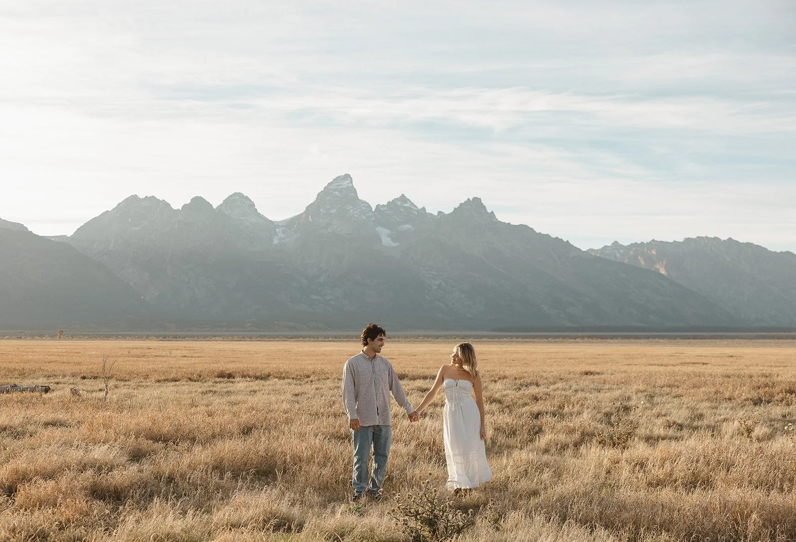 A man and woman holding hands in an open field with mountains in the background.