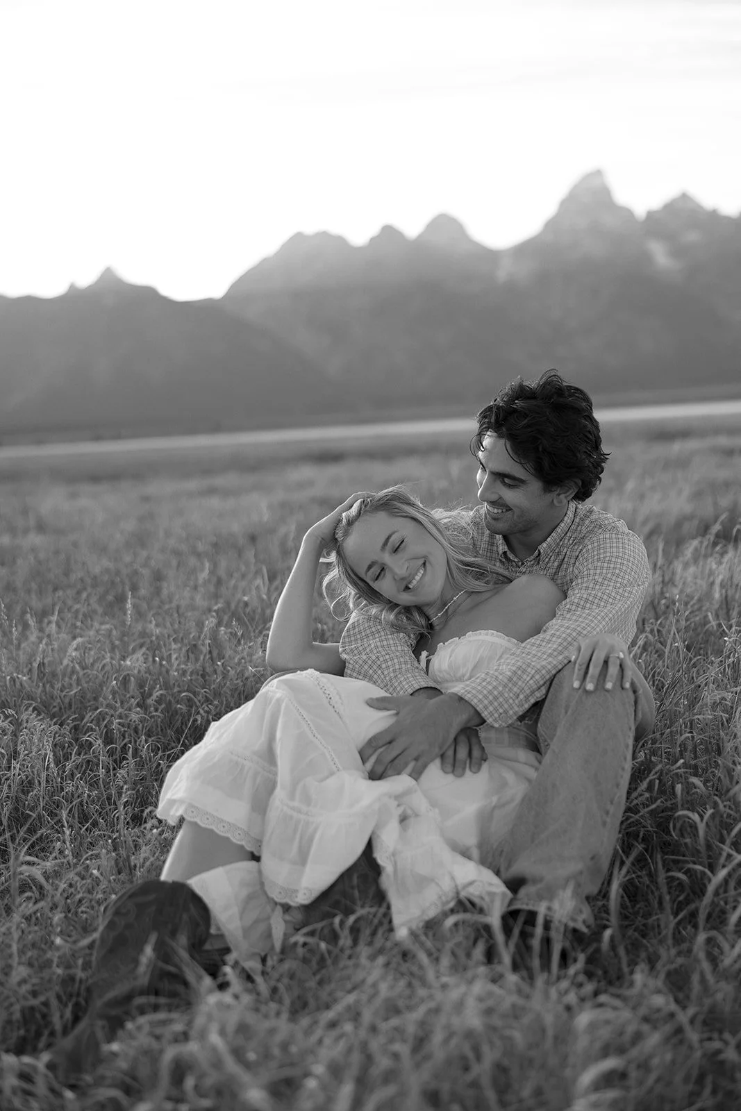 A black-and-white photo of a couple sitting in a grassy field with mountains in the background. They are smiling and embracing each other happily.