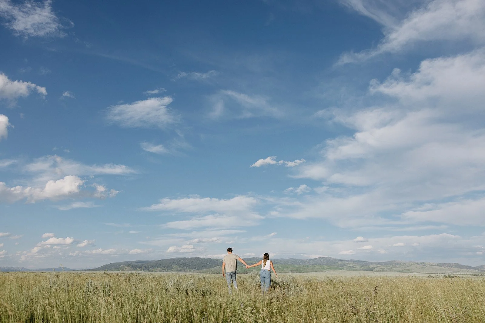 A man and woman walking hand in hand through a grassy field on a sunny day with a blue sky and scattered clouds.
