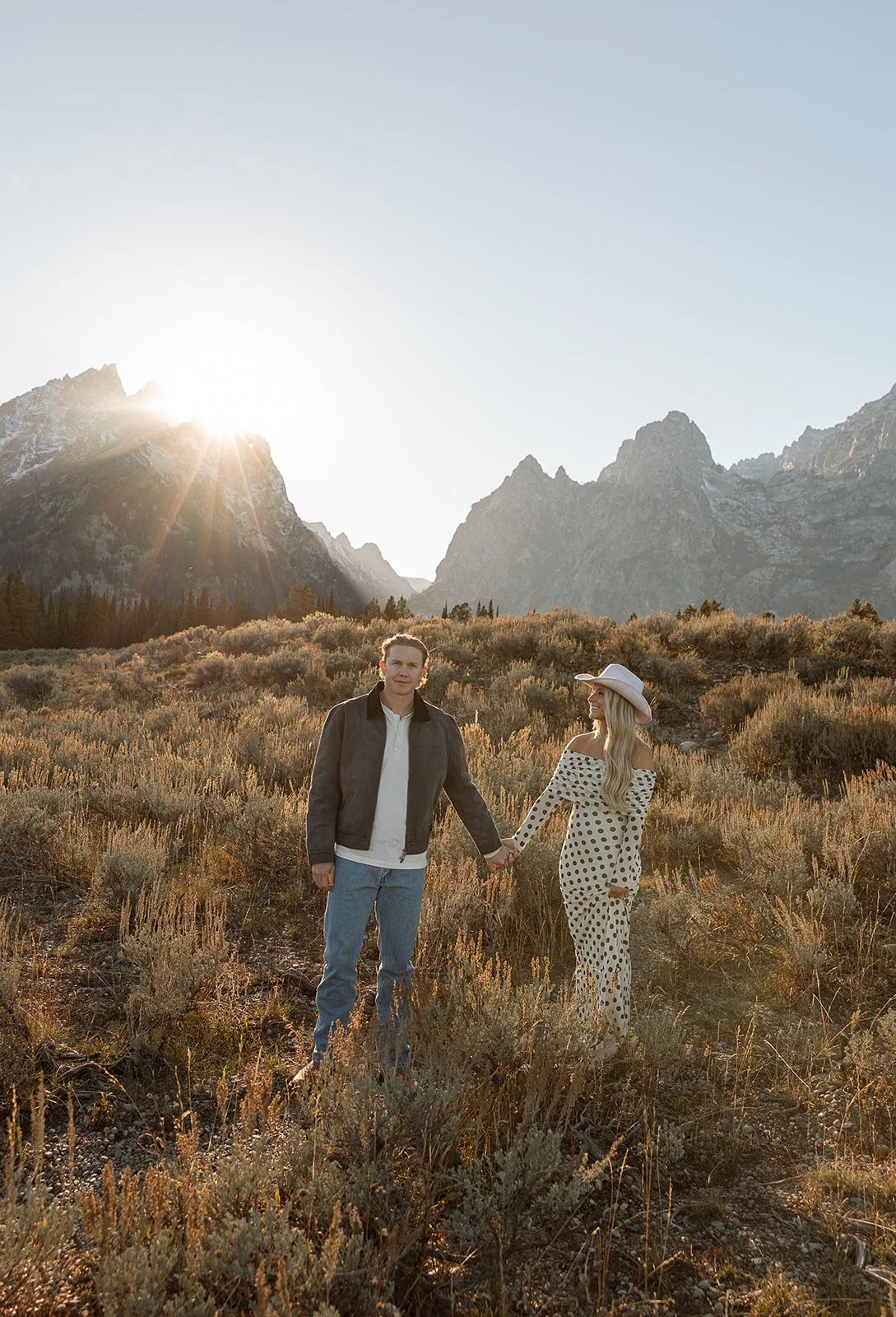A couple holding hands in a mountainous landscape at sunset or sunrise, with tall mountains and rugged terrain in the background.