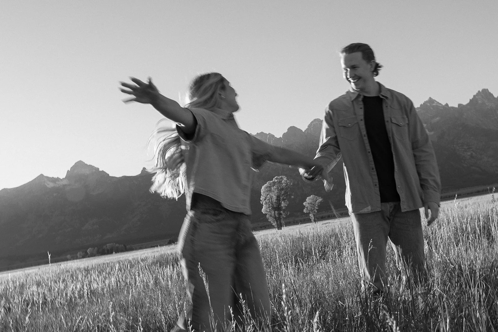 A black and white photo of a smiling couple holding hands in a field with mountains in the background, one person with arms outstretched.