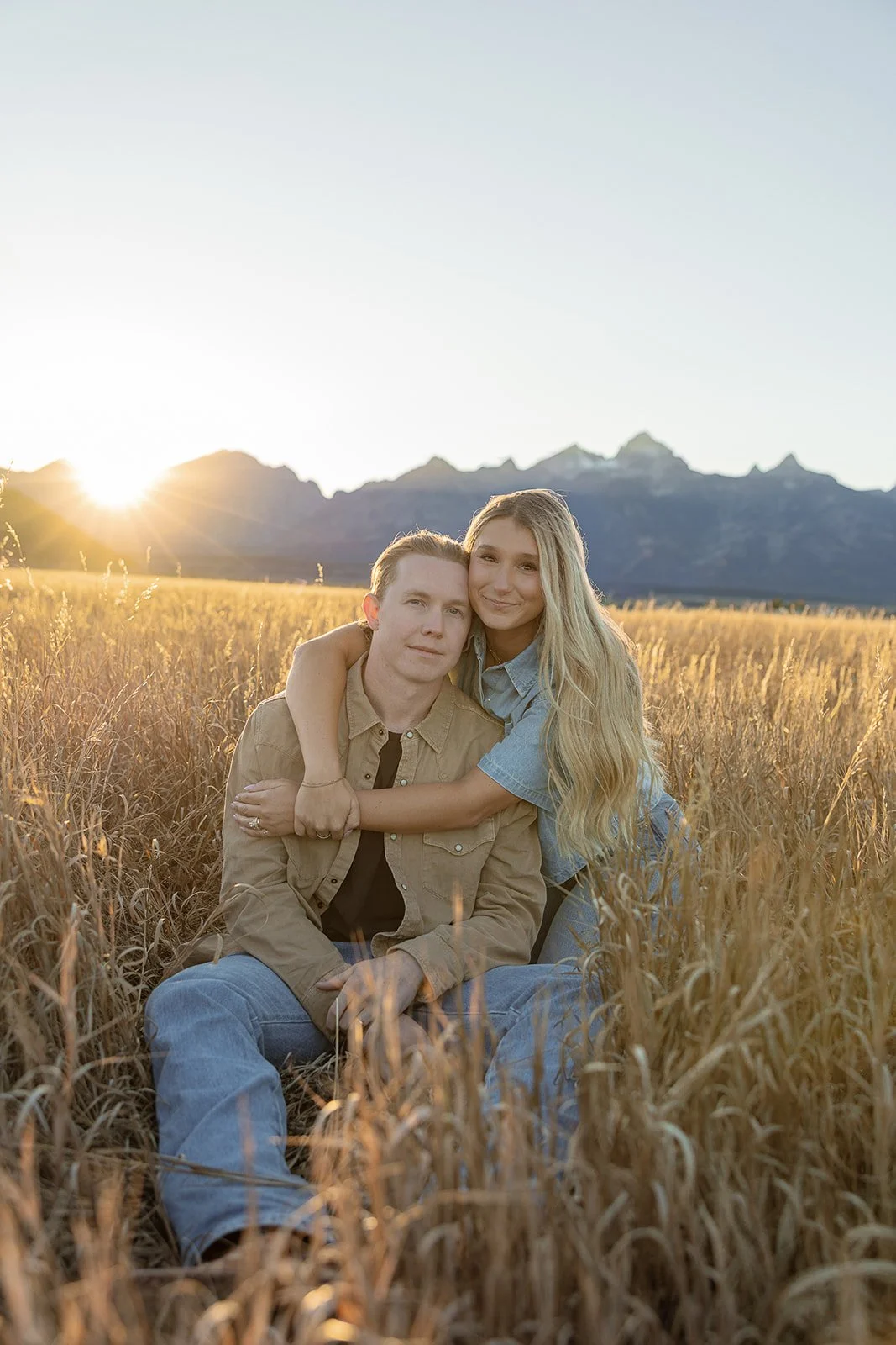 A couple sitting in a golden field at sunset with mountains in the background, embracing each other.