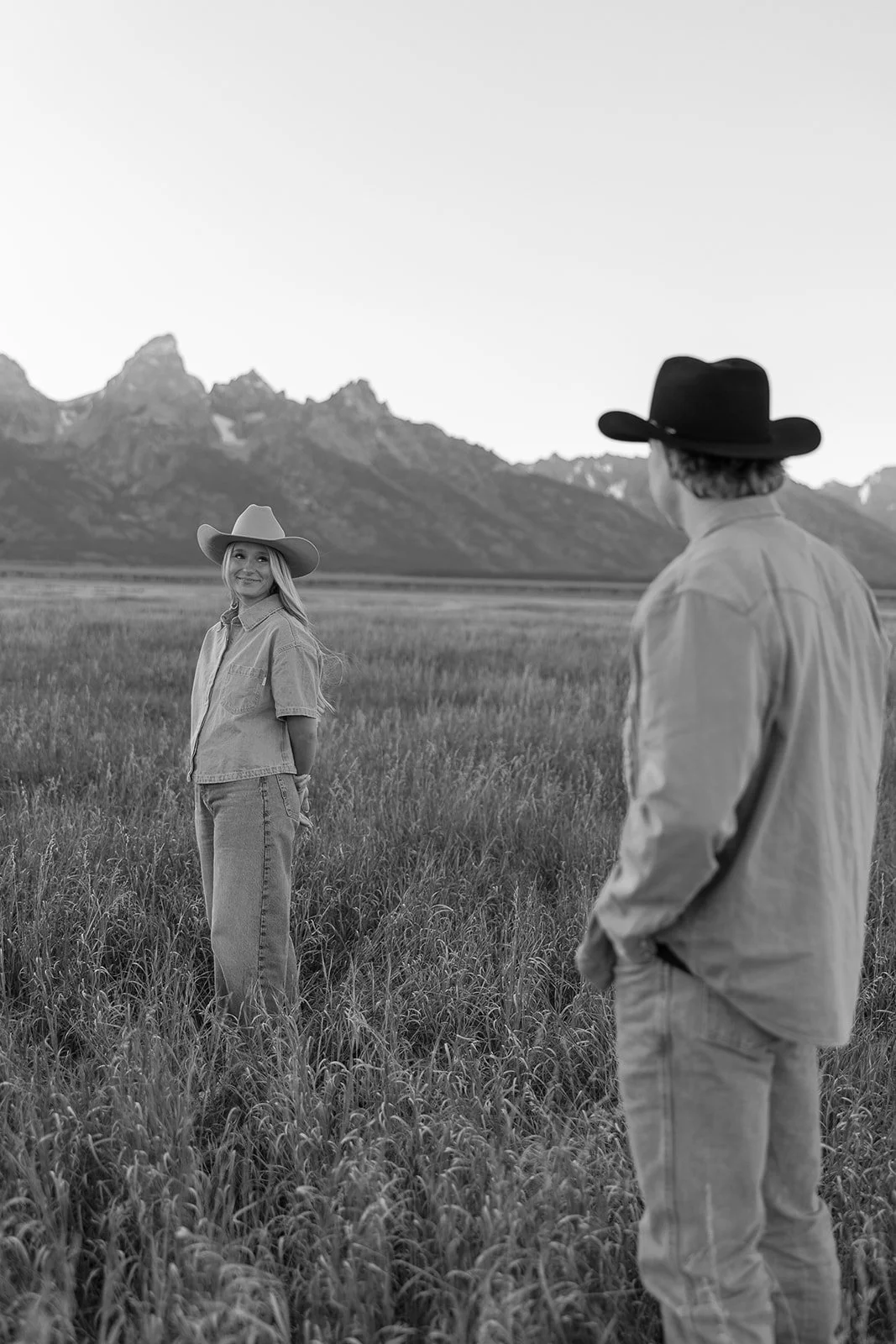 A black and white photo of a man and woman in cowboy hats standing in a grassy field with mountains in the background. The woman is smiling at the camera while the man faces her.