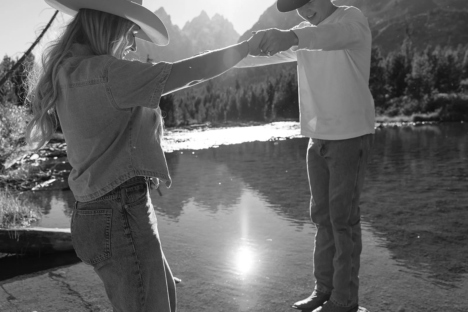 A woman and a young boy are holding hands and balancing on a riverbank near a river, with mountains and trees in the background, backlit by the sun.