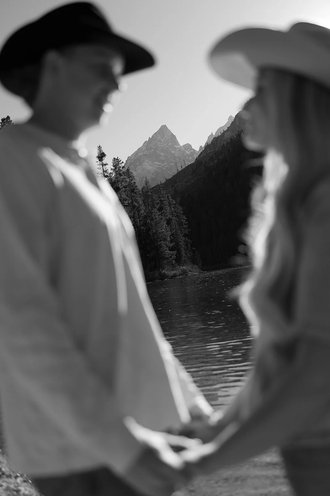 Two women holding hands by a river with mountains in the background, black and white image.