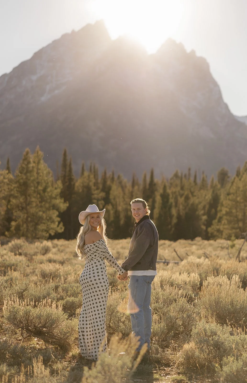 A couple holding hands and smiling in a field with mountain and pine forest background during sunset.