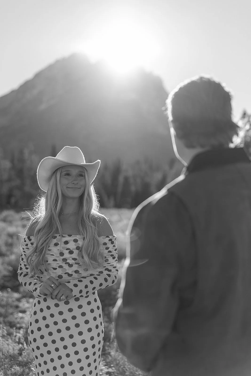 A woman in a polka dot dress and cowboy hat smiling at a man during sunset in a desert landscape.