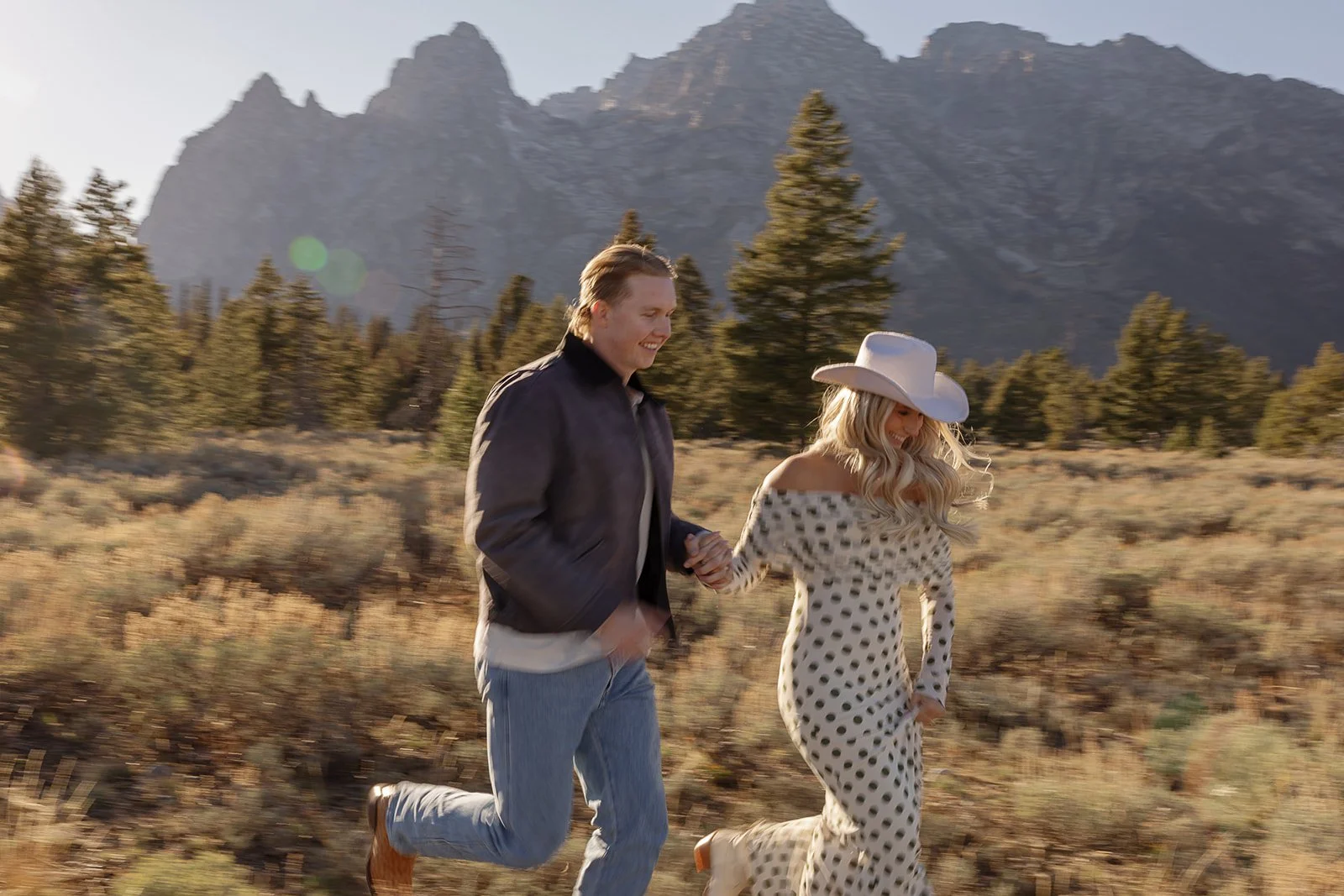 A man and woman are running hand in hand through a scenic outdoor area with mountains and pine trees in the background. The woman is wearing a wide-brimmed white hat and a polka dot dress, while the man is dressed casually in a jacket and jeans.