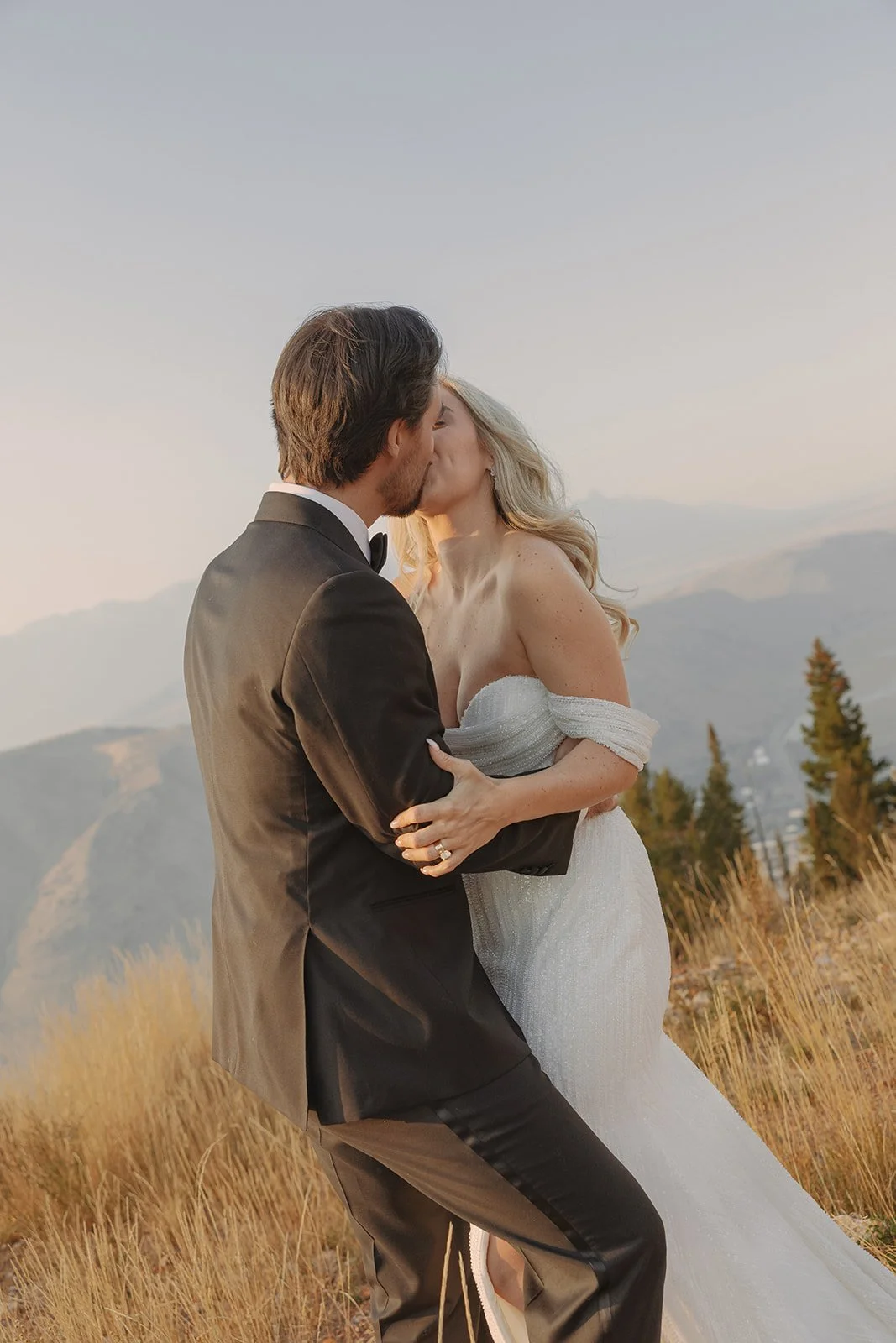 A bride and groom kissing outdoors during sunset, with mountainous landscape and trees in the background.