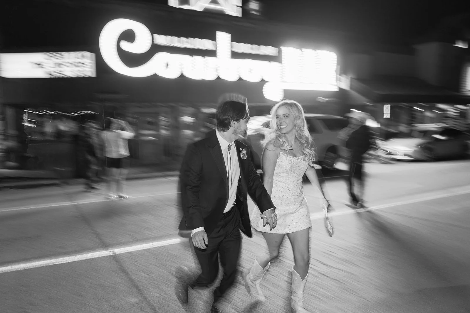 A black-and-white photo of a couple walking hand-in-hand on a city street at night, surrounded by blurred pedestrians and vehicles, with a brightly lit sign in the background.
