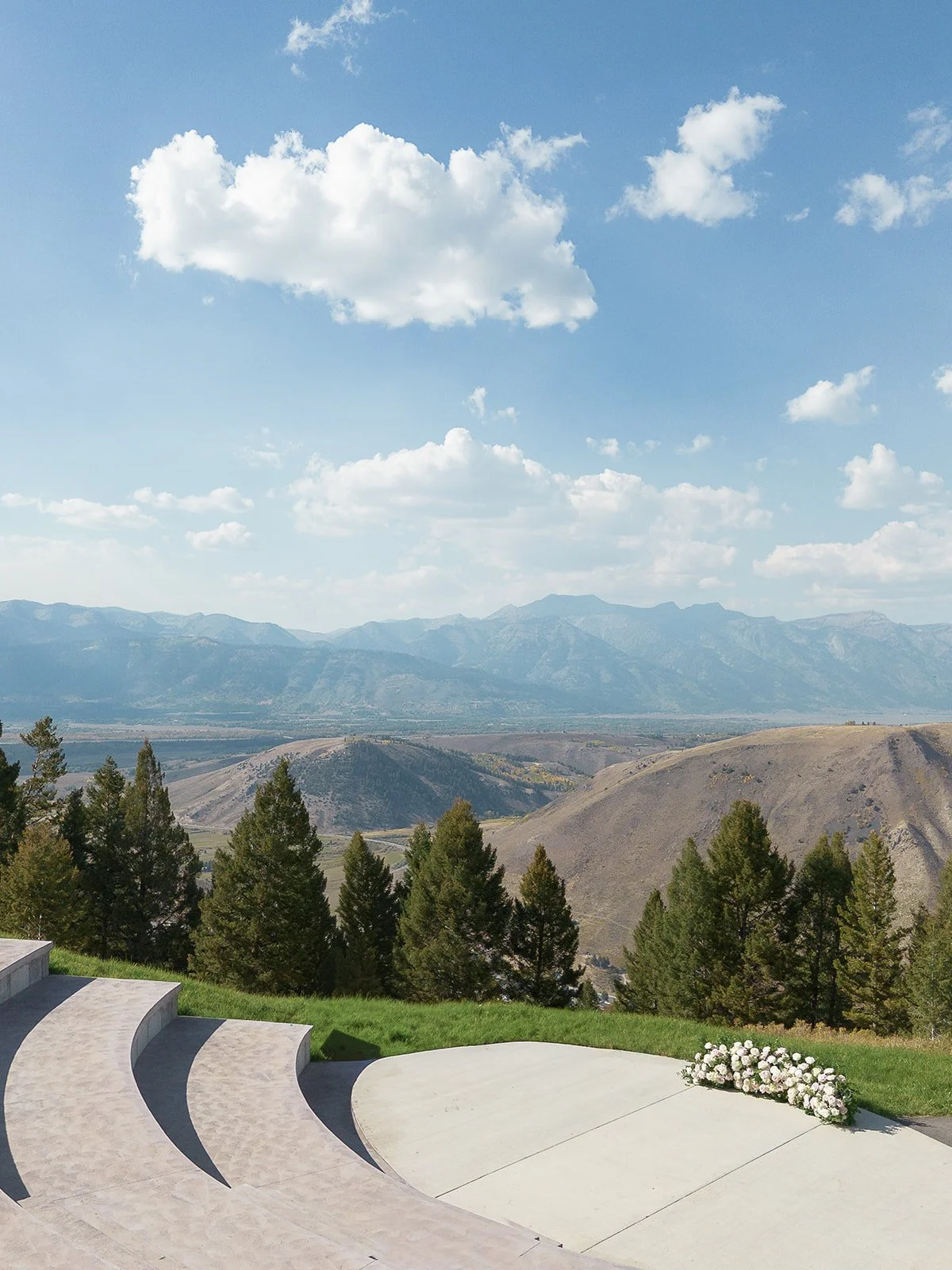 This image features a scenic view of mountains and a valley with partly cloudy skies. In the foreground, there is a curved concrete platform with steps and a floral arrangement on the ground.