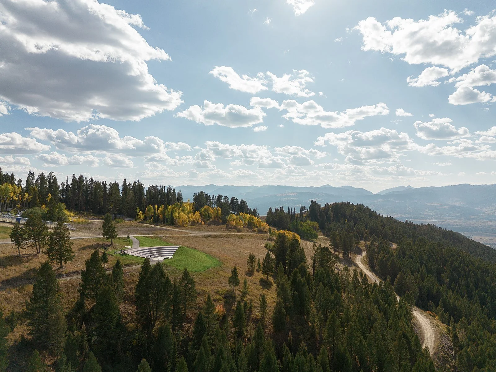 A scenic view of a mountainous landscape with a mix of green and yellow trees, winding dirt roads, and a partly cloudy sky.