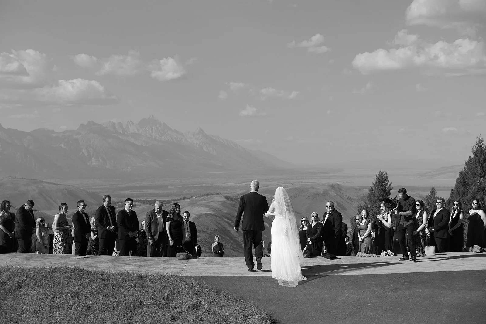 A man and woman in wedding attire walk hand in hand towards a gathering of people on a lawn overlooking mountains and a valley.