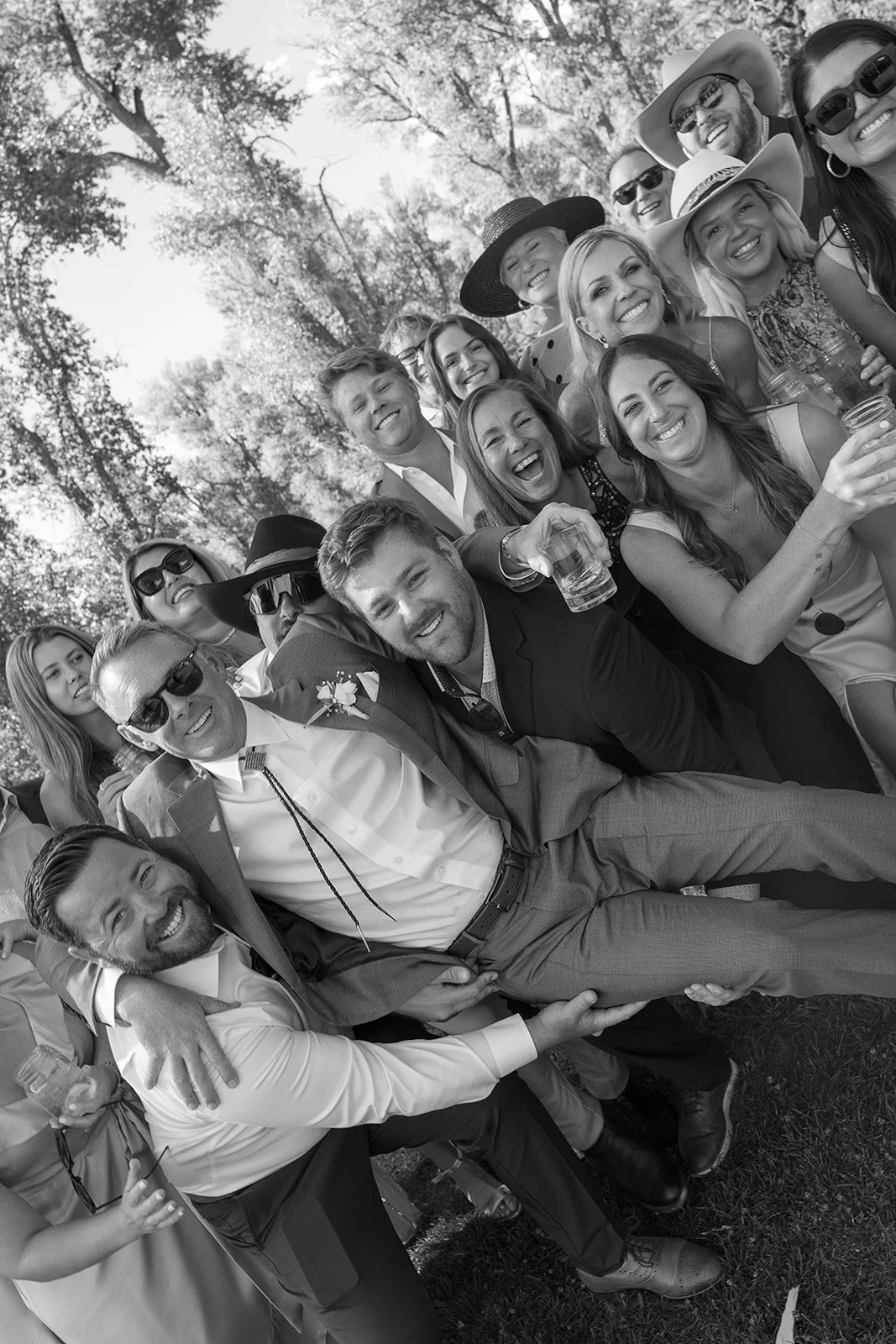 A group of people gathered outdoors for a celebration, smiling and laughing, with some holding drinks. The photo is in black and white and appears to be taken during a sunny day with trees in the background.