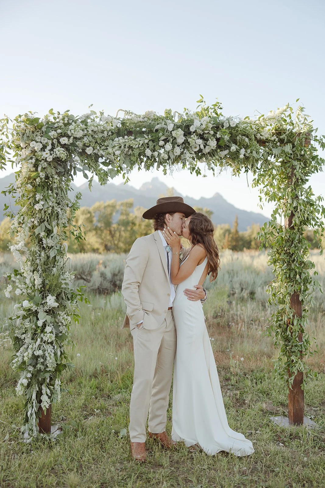 A couple dressed in wedding attire sharing a kiss under a floral arch outdoors with mountains in the background.