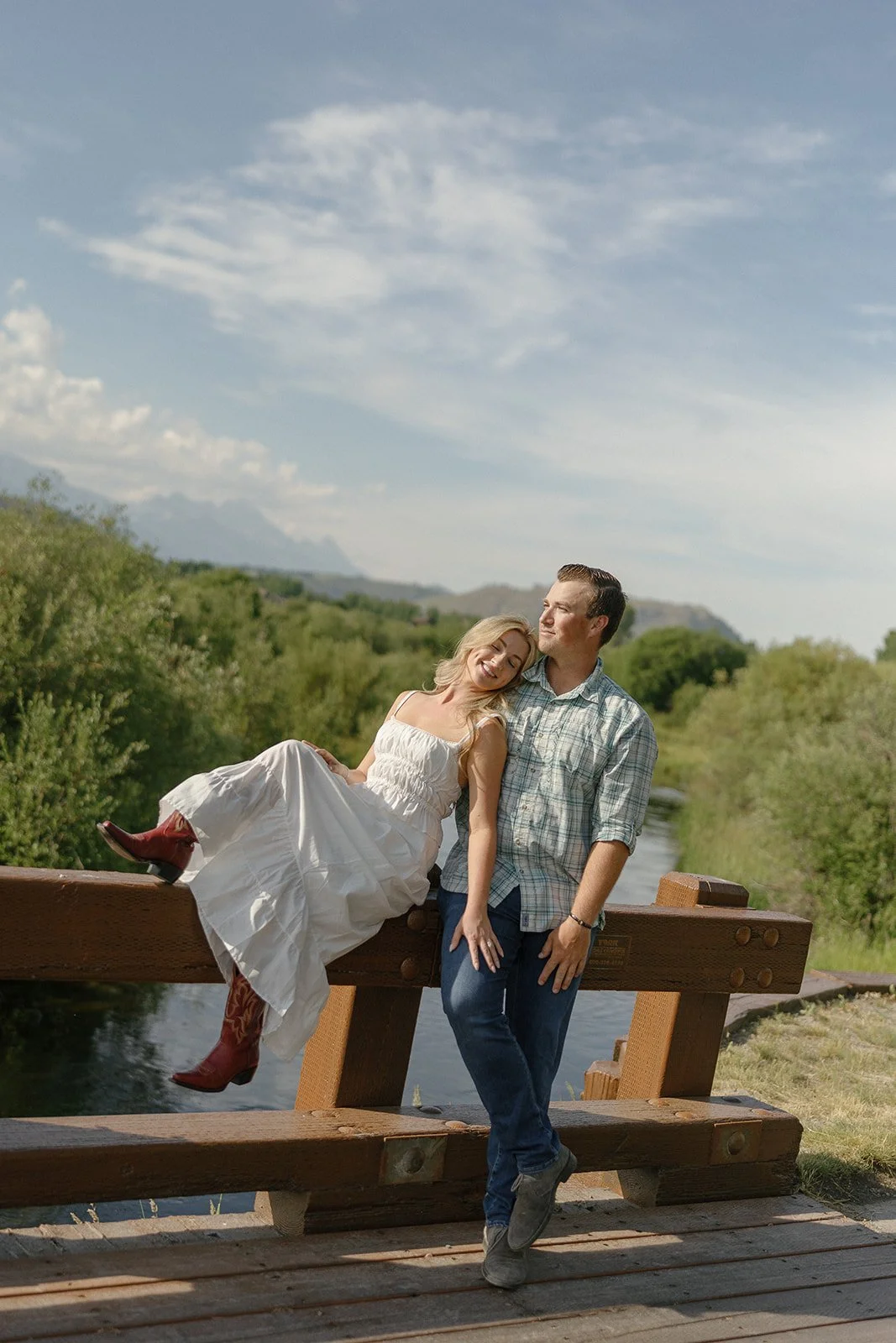A smiling woman in a white dress and cowboy boots leaning against a man in a plaid shirt, standing on a wooden bridge over a river in a lush green landscape with mountains in the background.