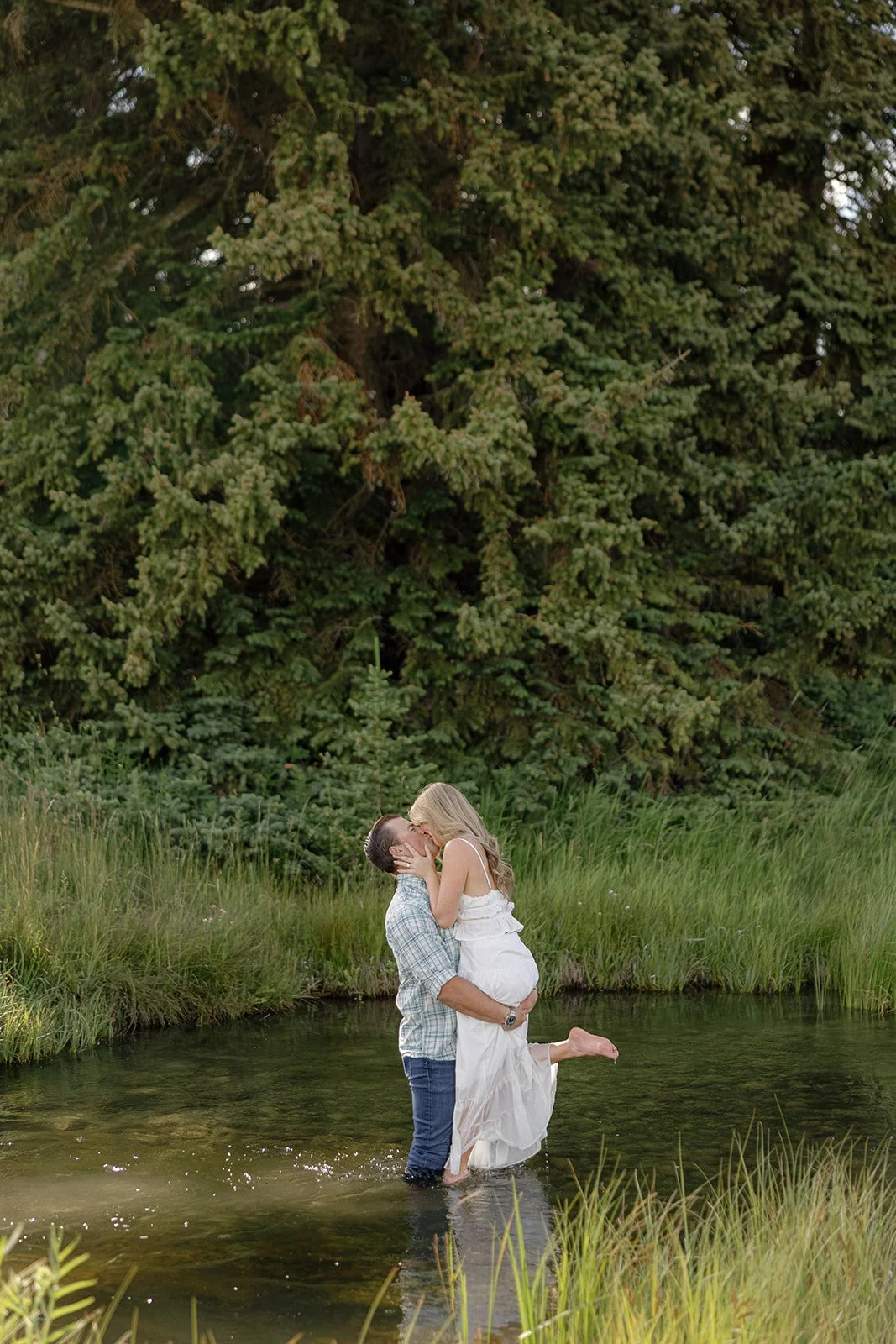 A couple stands in a shallow creek surrounded by green grass and trees. The man lifts the woman in his arms as they kiss.