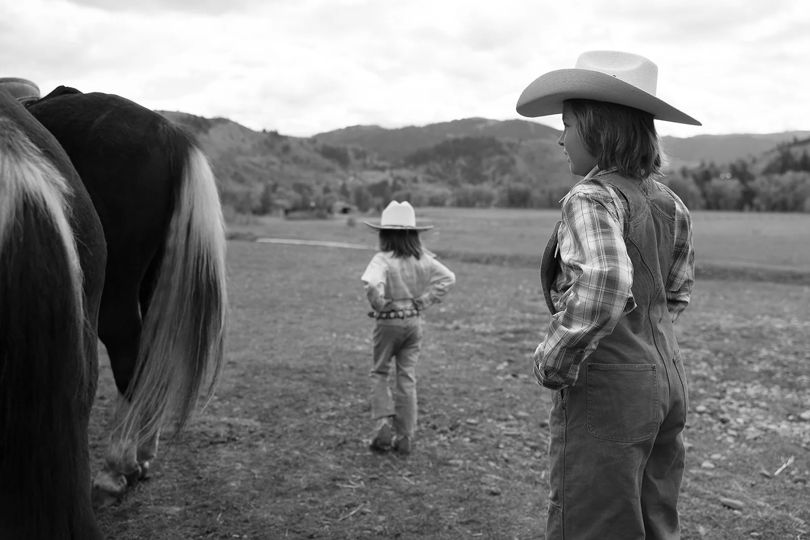 Two young girls wearing cowboy hats and plaid shirts stand in a rural outdoor landscape with rolling hills in the background. One girl is close to the camera, looking at horses nearby, while the other girl stands further away with hands on hips.