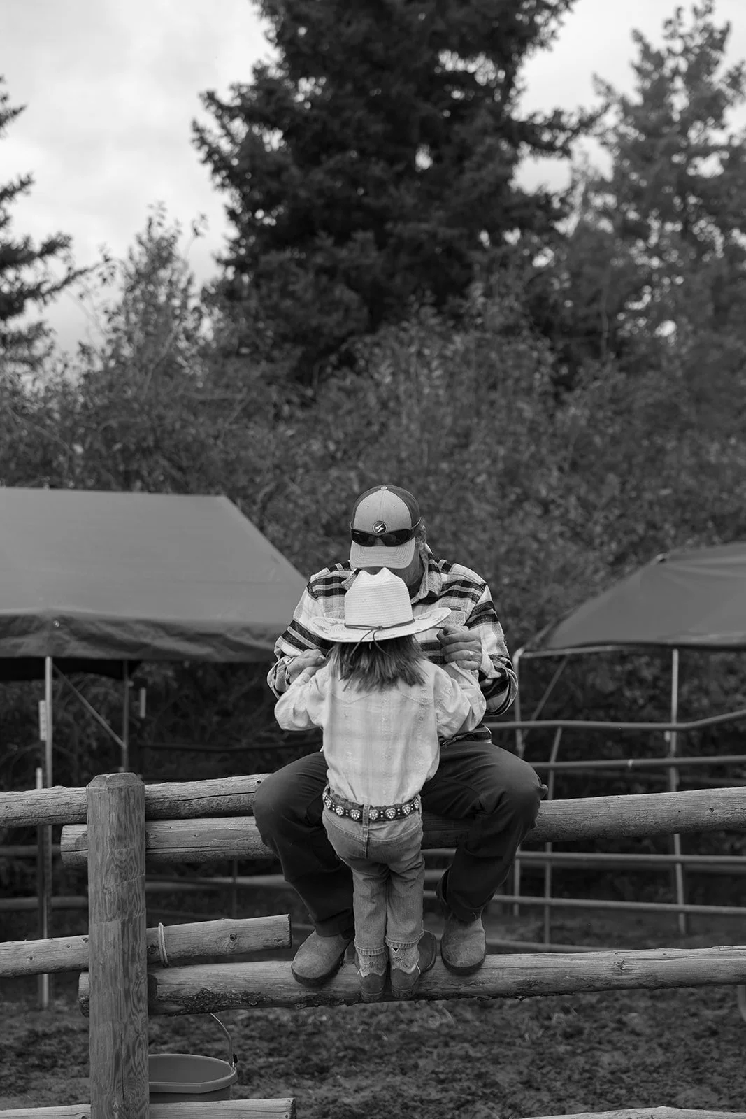 An adult man is kneeling on a wooden fence, presenting a small object to a young girl. The girl is wearing a cowboy hat, jeans, and a long-sleeve shirt, while the man is wearing a baseball cap, a plaid shirt, and boots. They are outdoors, with trees 