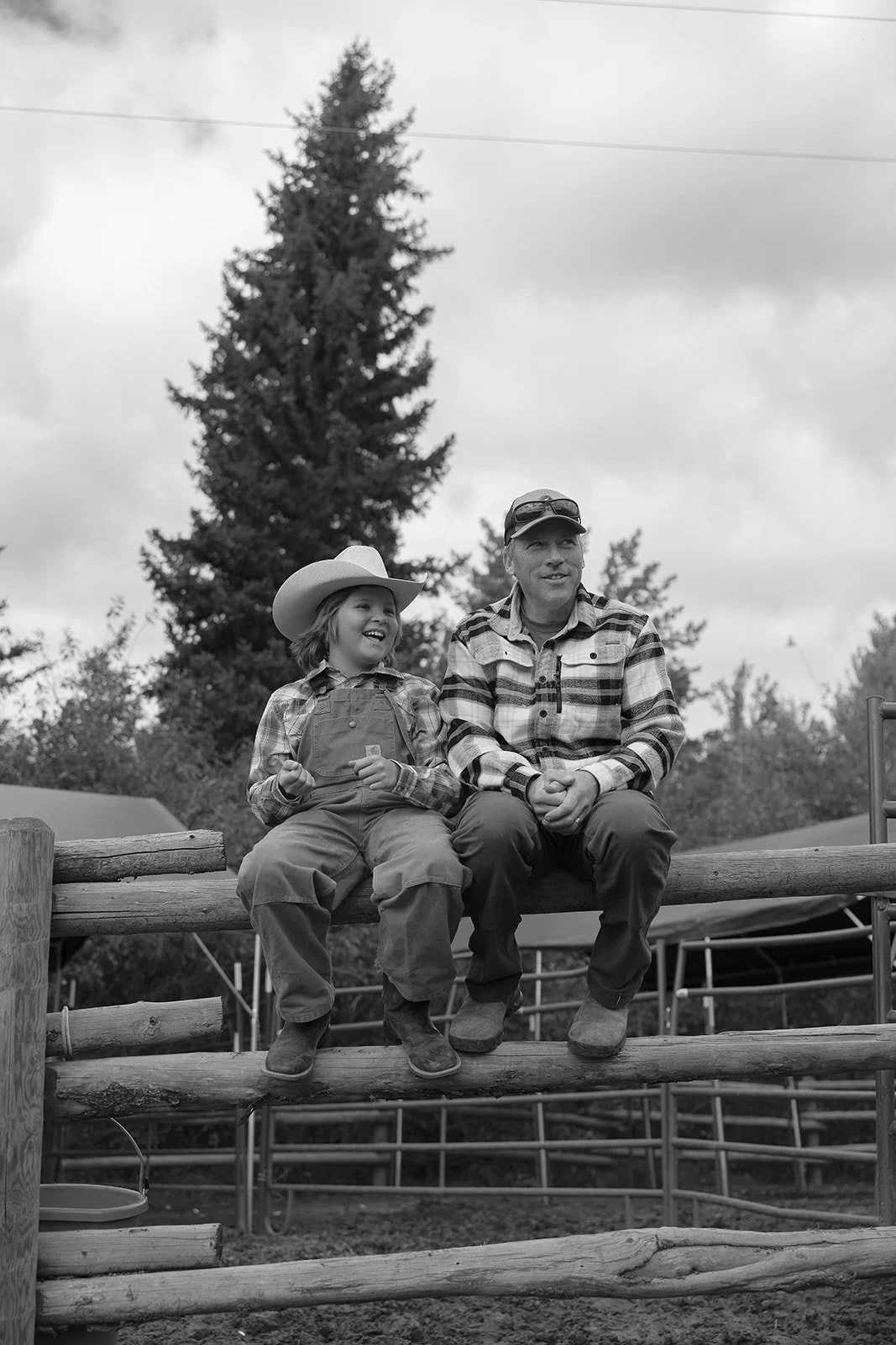 A man and a young girl sitting together on a wooden fence outdoors, both smiling. The girl is wearing a cowboy hat and overalls, while the man is in a plaid shirt and cap. There are trees and a cloudy sky in the background.