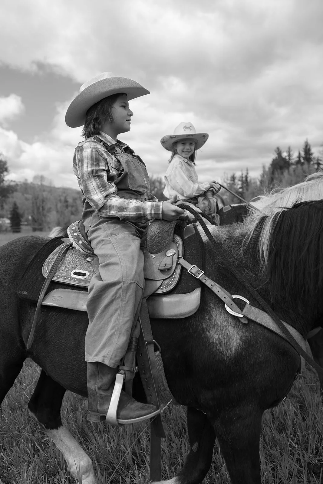 Black and white photo of two children riding horses outdoors, wearing cowboy hats and western clothing, with a scenic background of trees and clouds.