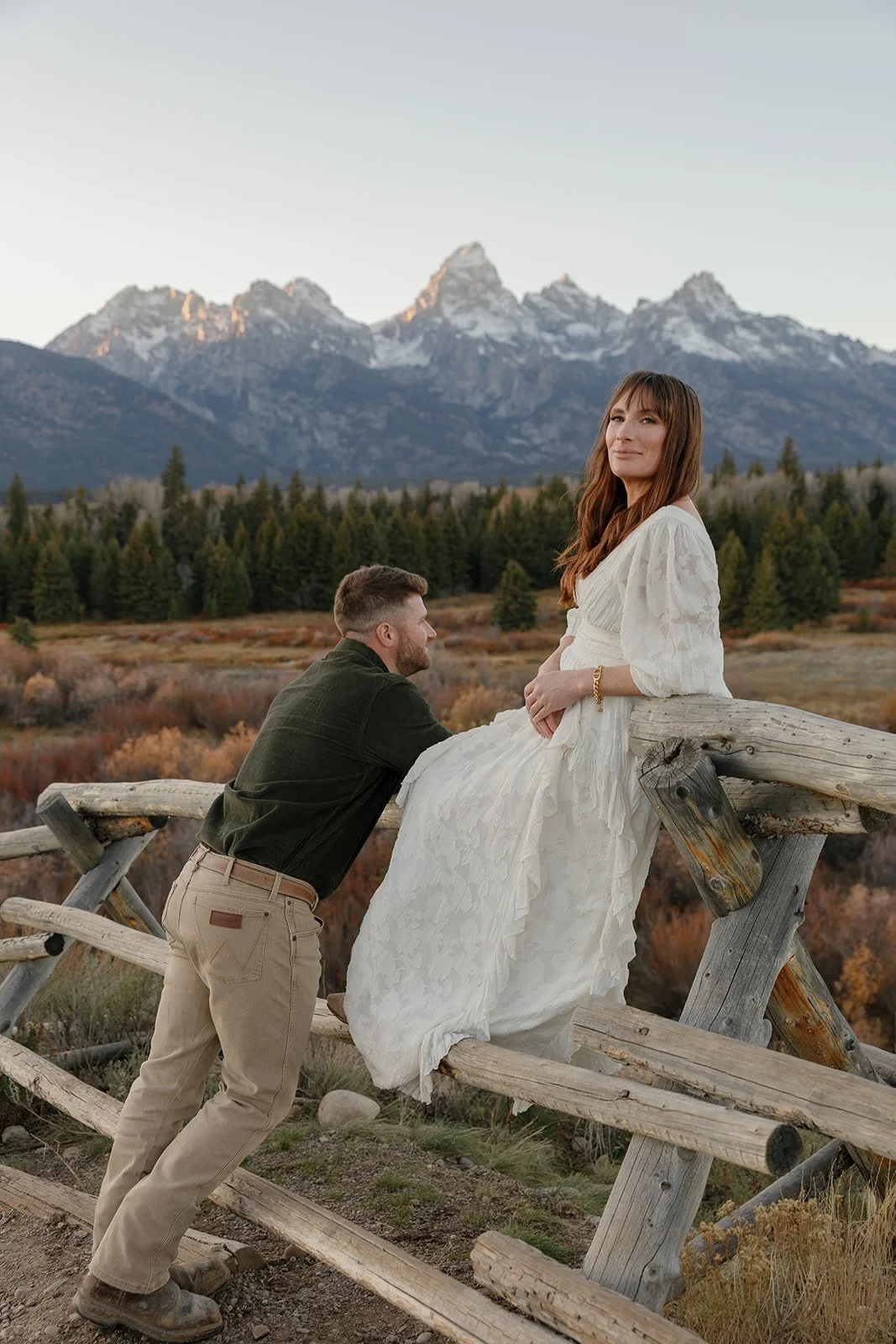 A man kneeling on a rustic wooden fence, gazing at a woman sitting on the fence, with mountain peaks and a forest in the background.