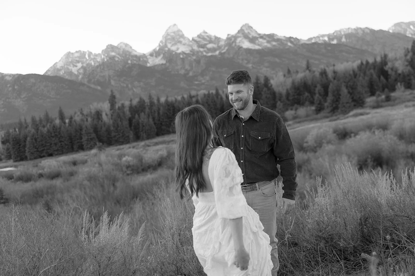 A man and woman standing in a field with mountains in the background, smiling at each other.