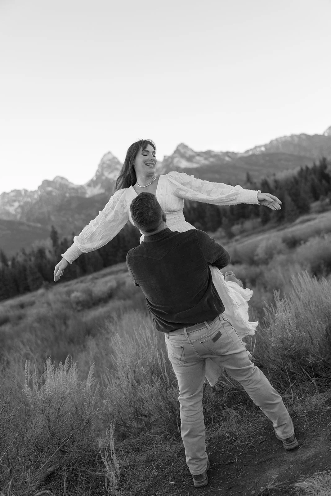A couple outdoors, with the man lifting the woman in a field, mountains in the background, black and white photo.