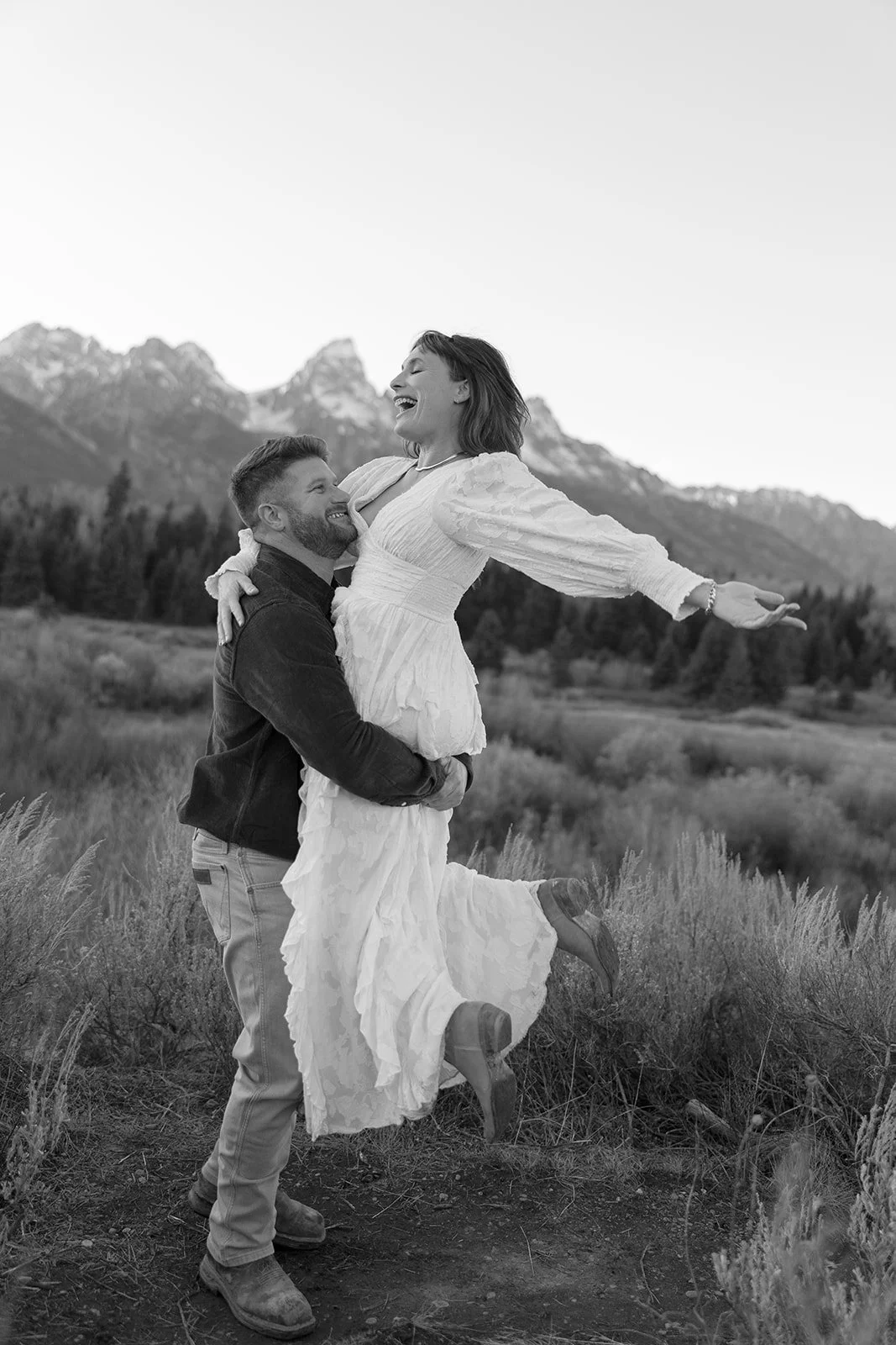 A man lifting a woman in a field with mountains in the background, both laughing joyfully.