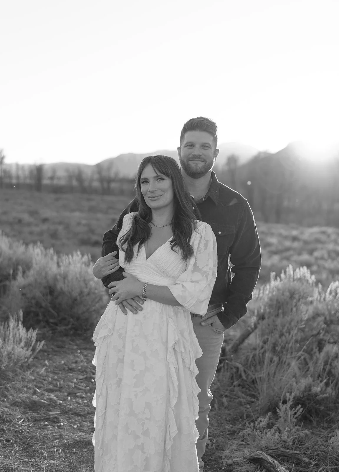 A black and white photo of a couple standing outdoors in a field with mountains in the background, at sunset or sunrise, with the woman in a white dress and the man in a dark shirt.