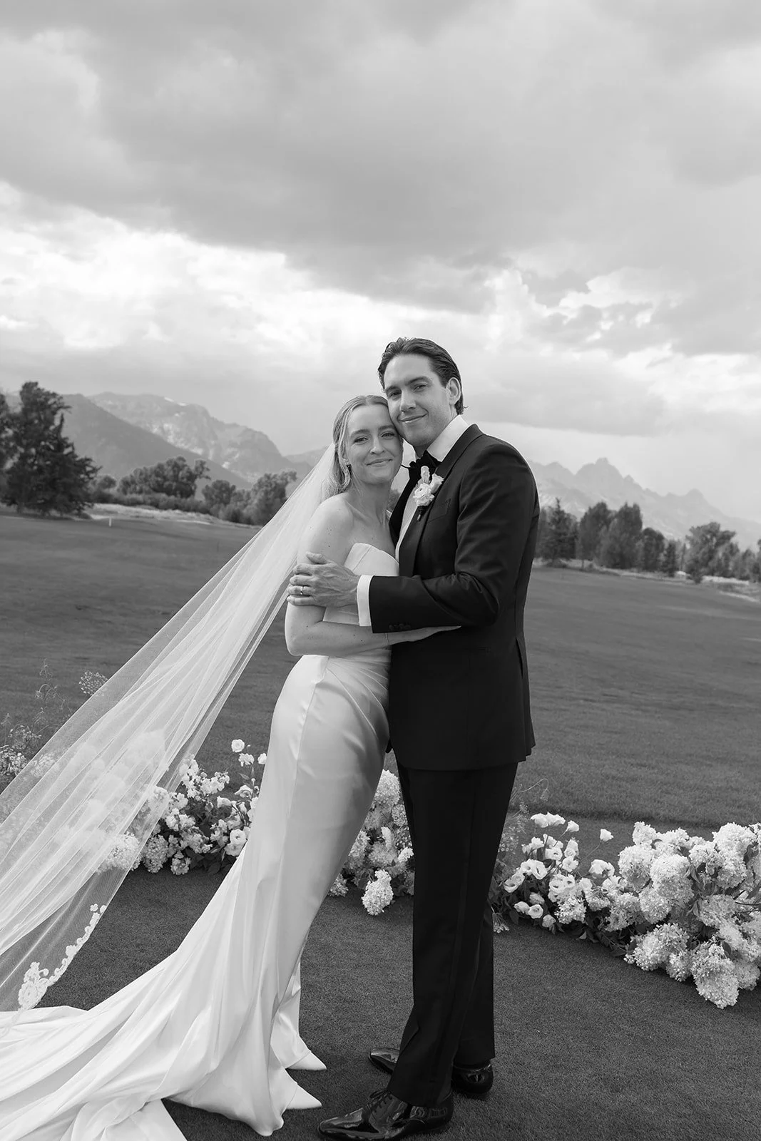 Black and white photo of a bride and groom hugging outdoors with mountains in the background, flowers on the ground, and a cloudy sky overhead.