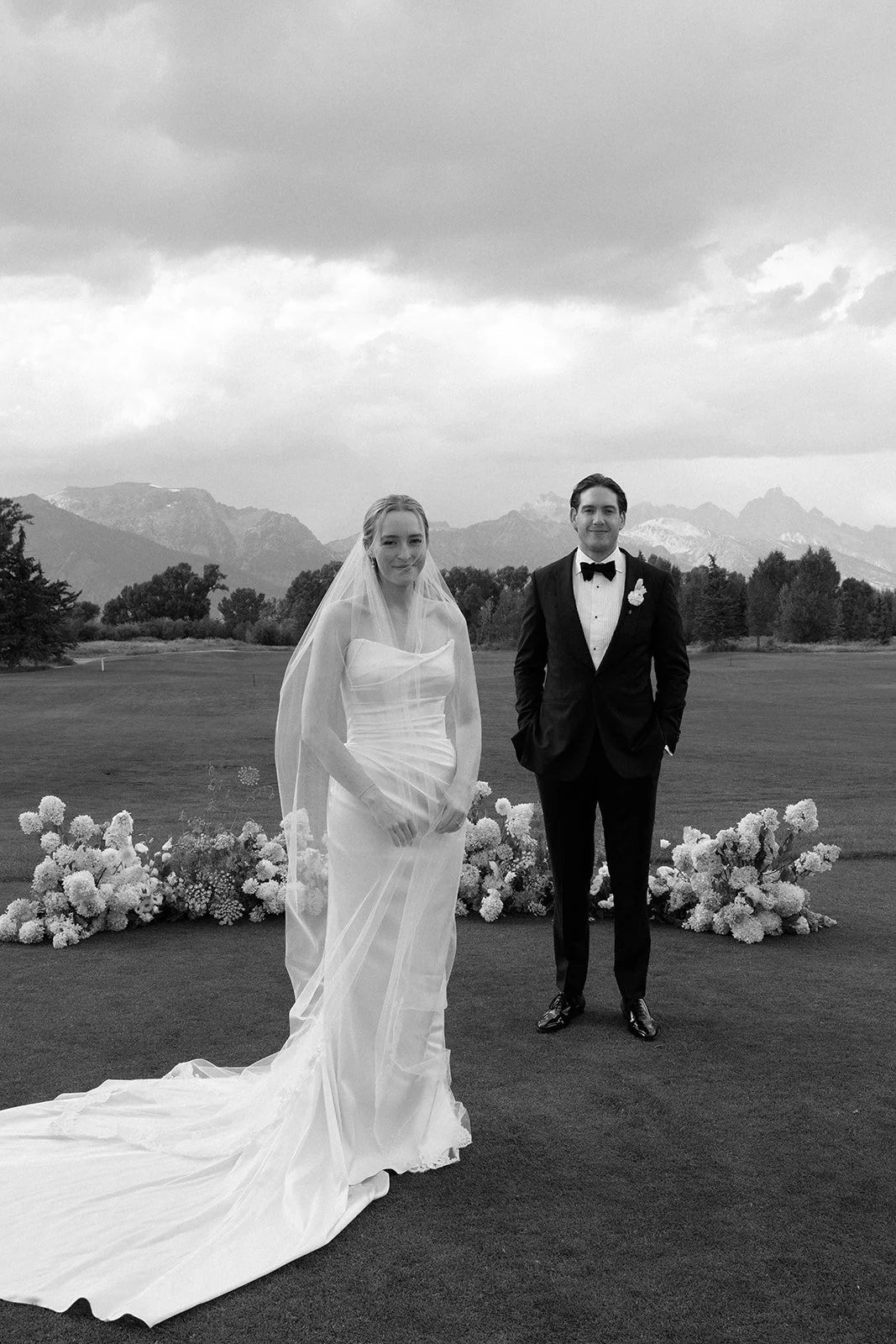 Black and white photo of a bride and groom standing outdoors on a grassy field, with mountains in the background, and floral arrangements behind them.