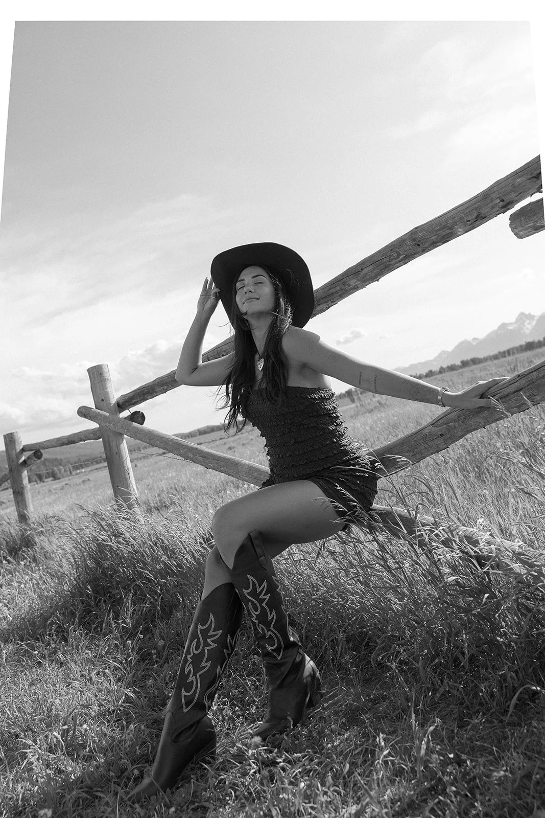 A woman wearing a large hat and a strapless dress, sitting on a wooden fence in an open grassy field, with mountains in the background, smiling with eyes closed.