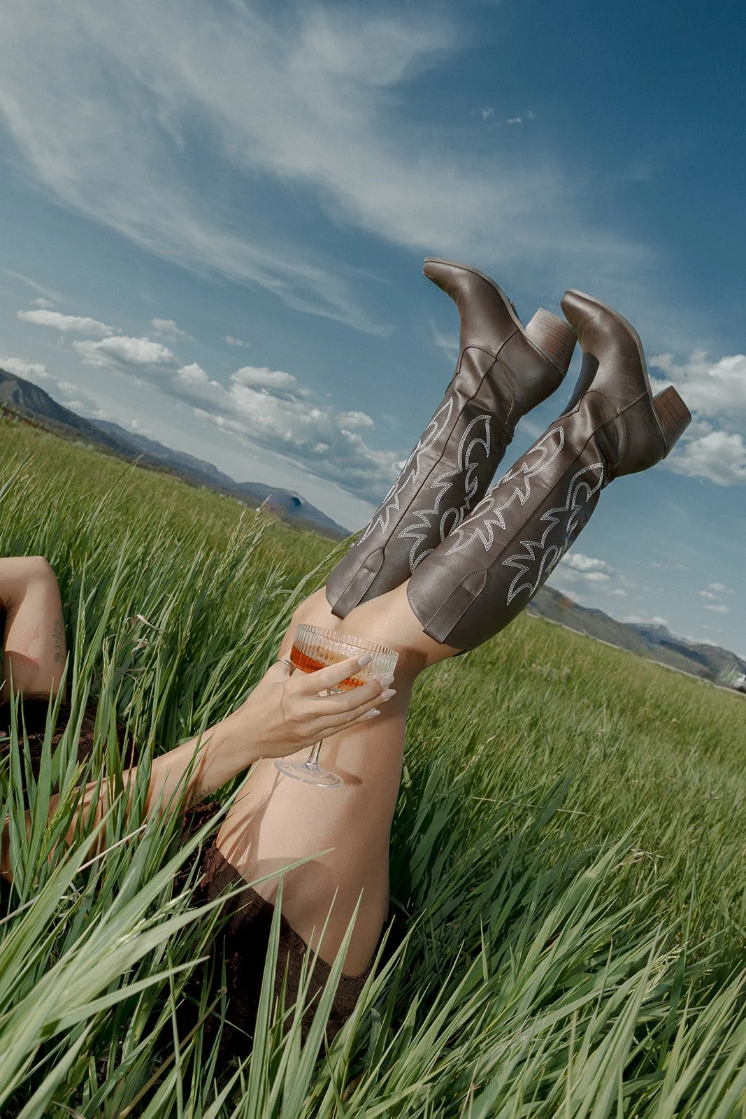 Person relaxing in a grassy field holding a cocktail, with tan cowboy boots with white decorative stitching, legs raised in the air, under a blue sky with clouds, mountains in the background.