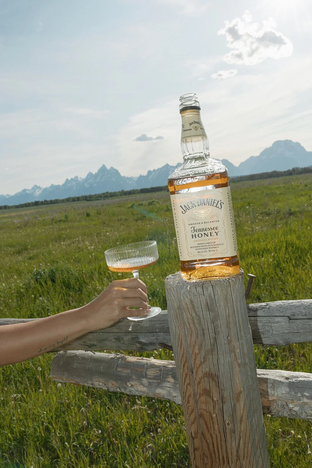 A hand holding a glass of whiskey with a bottle of Jack Daniel's Tennessee Honey whiskey on a wooden fence with a scenic mountain range in the background.