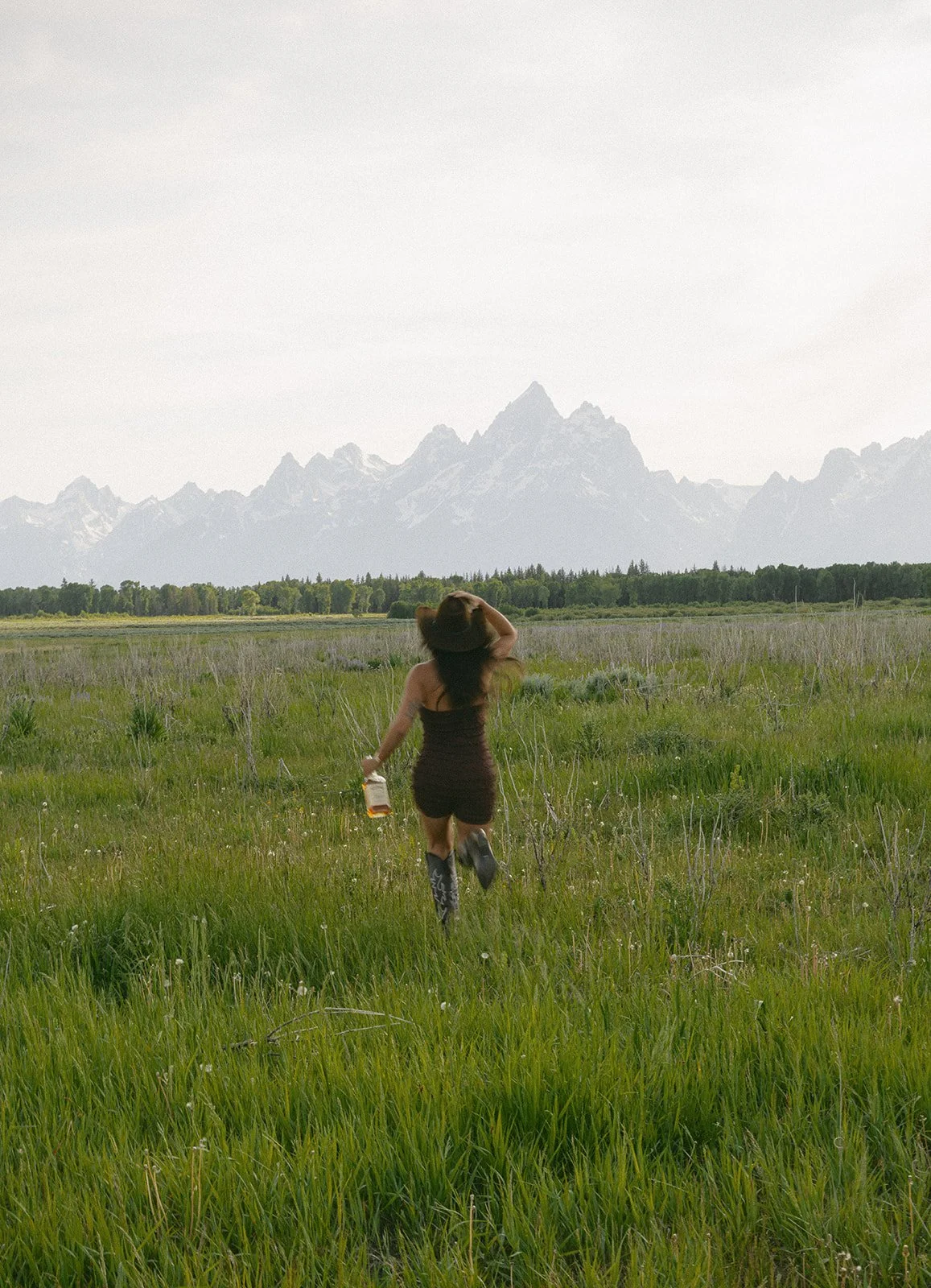 A woman in a black dress and cowboy boots running through a grassy field with mountains in the background.