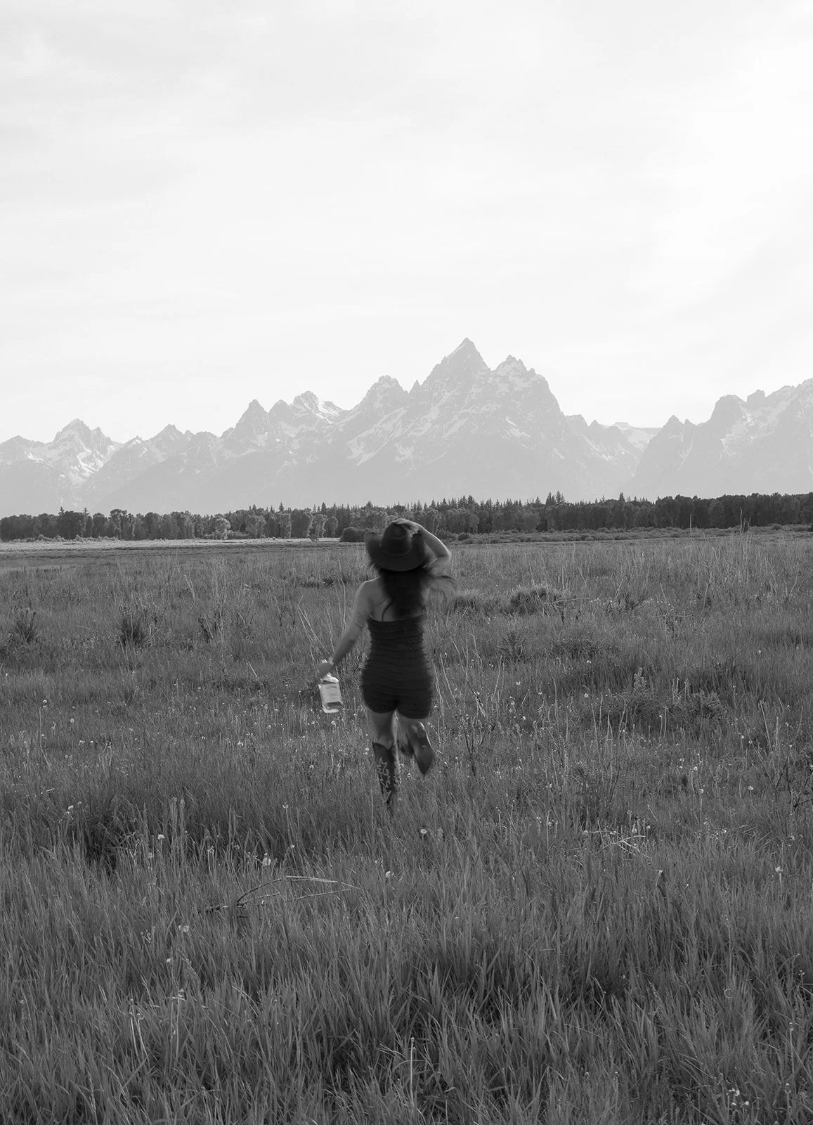 A woman wearing a hat and dress holding a drink walking through a grassy field with mountains in the background.