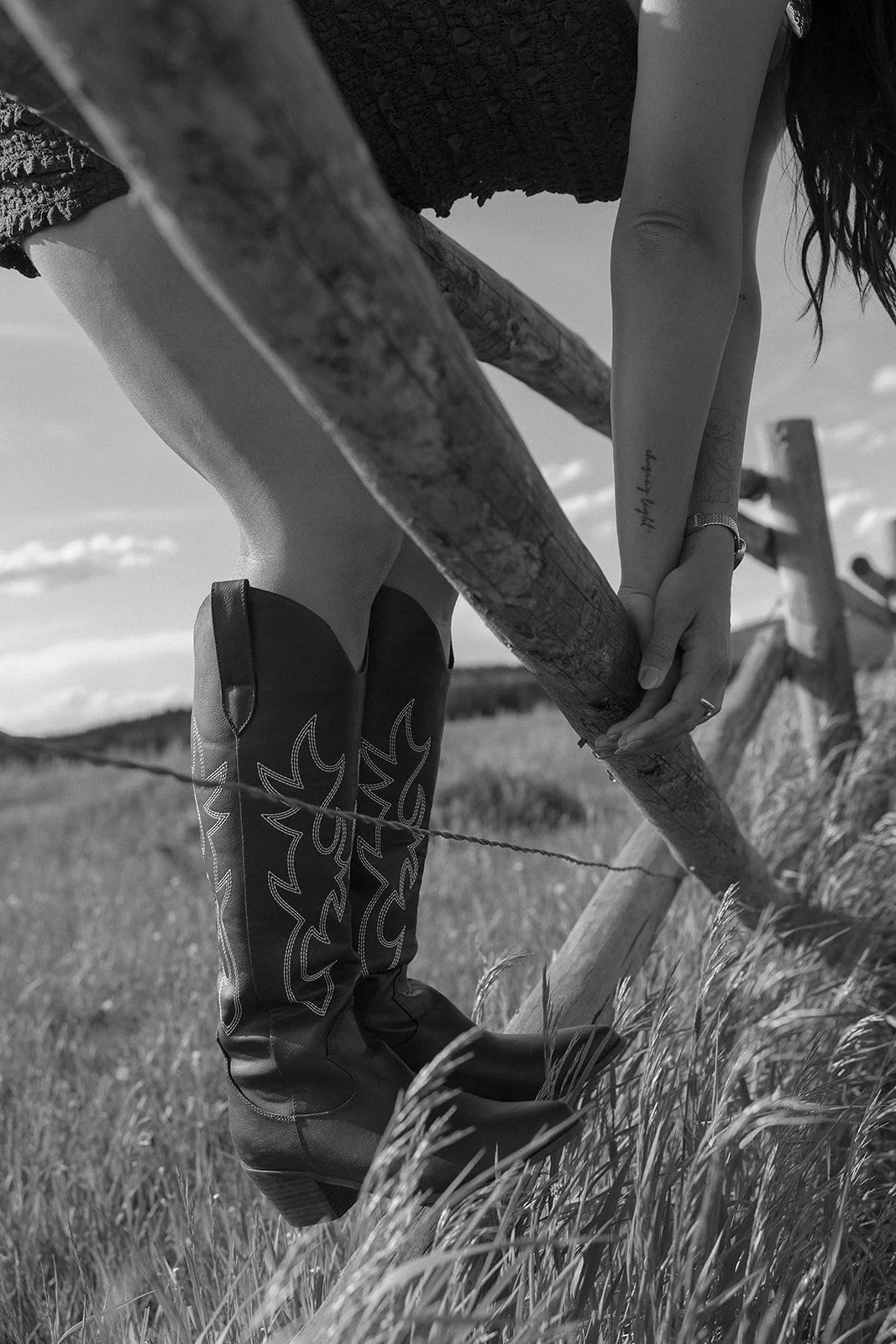 A woman wearing cowboy boots and a dress, holding onto a wooden fence in a grassy field with a cloudy sky.