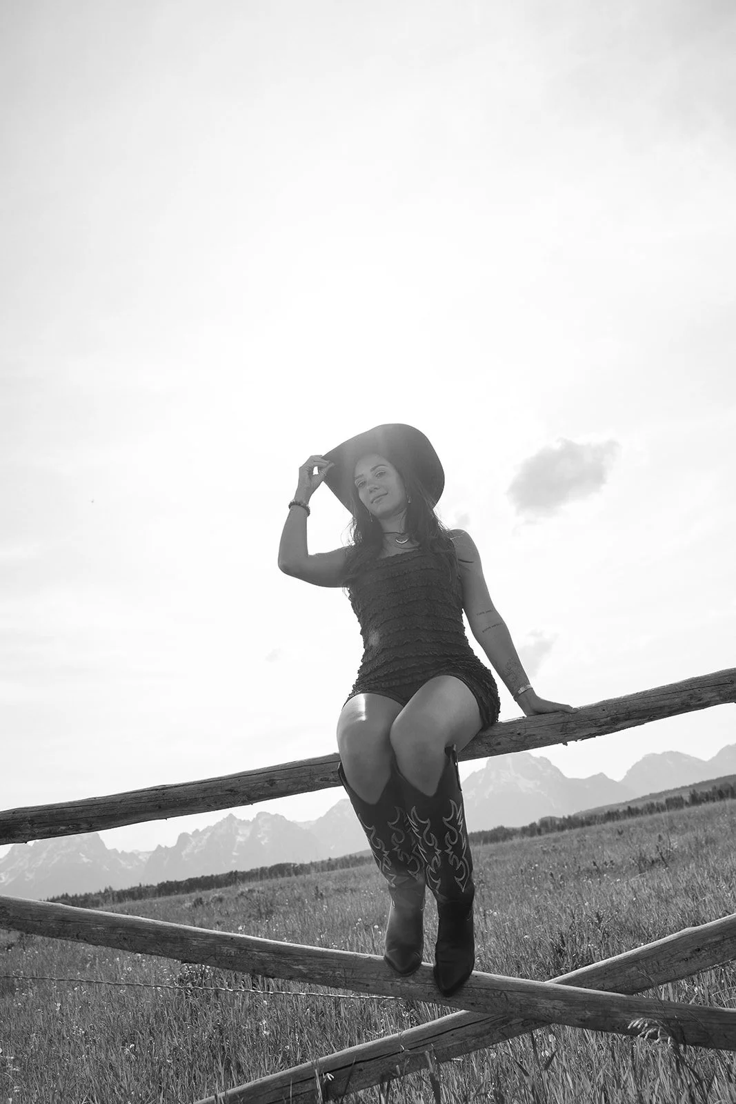 A woman sitting on a wooden fence in a field with mountains in the background, wearing a hat, dress, and cowboy boots, smiling at the camera.
