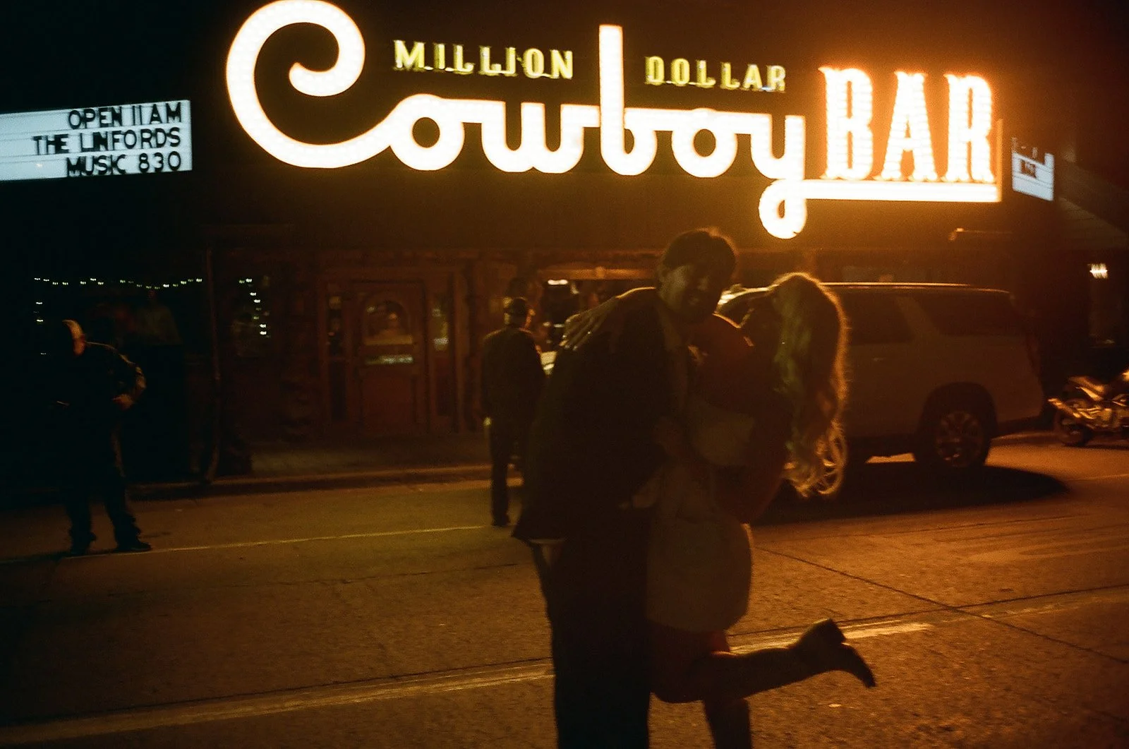 Couple dancing in front of a neon-lit Cowboy Bar sign at night with other people, cars, and motorcycles visible in the background.