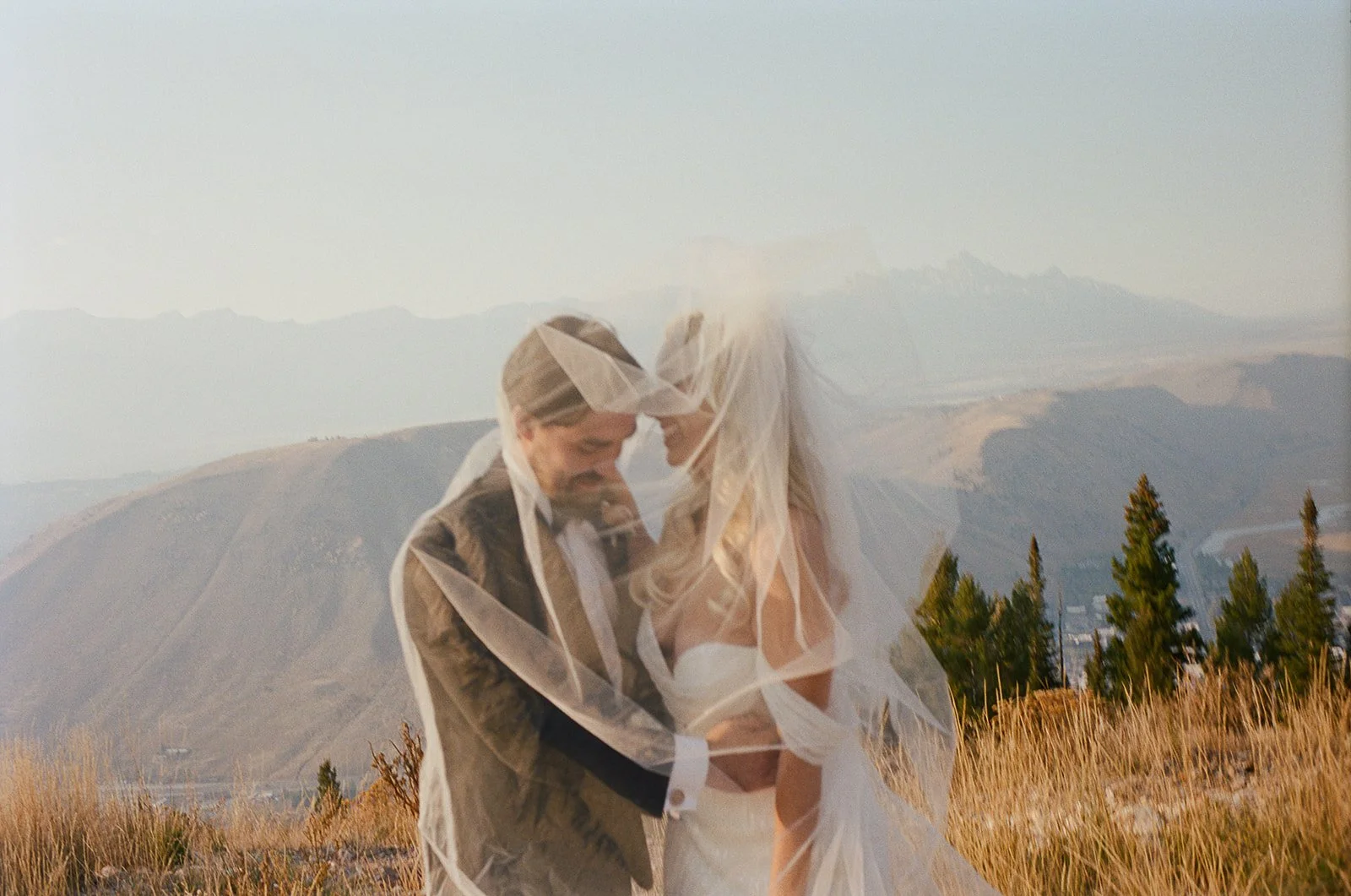 A bride and groom standing close together, smiling under a wedding veil outdoors with mountains and trees in the background.