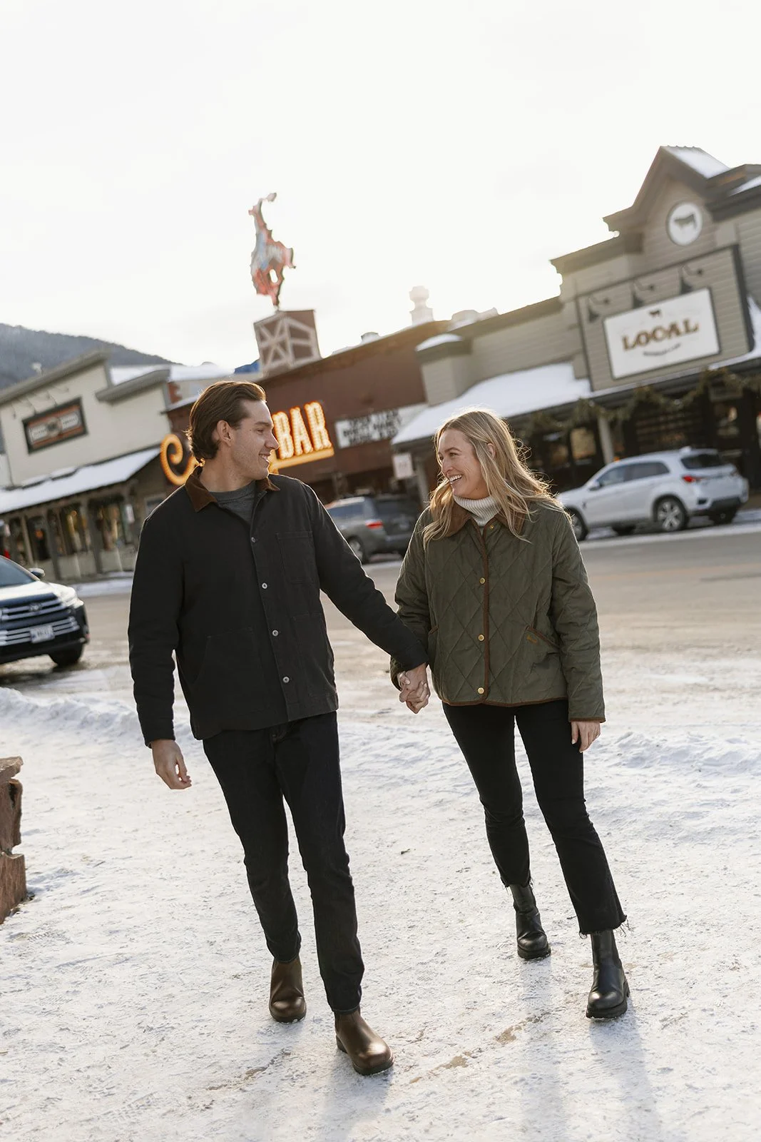 A young couple walking hand in hand on a snowy street in a small town, smiling at each other, with shops and cars in the background.