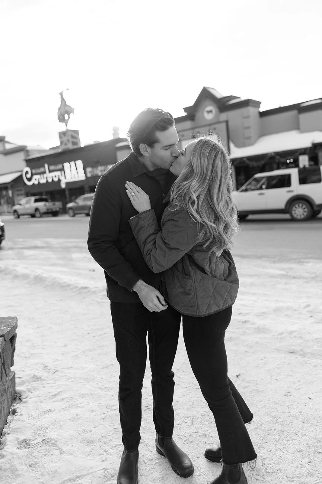 A black and white photo of a young couple kissing outdoors on a beach with stores and cars in the background.