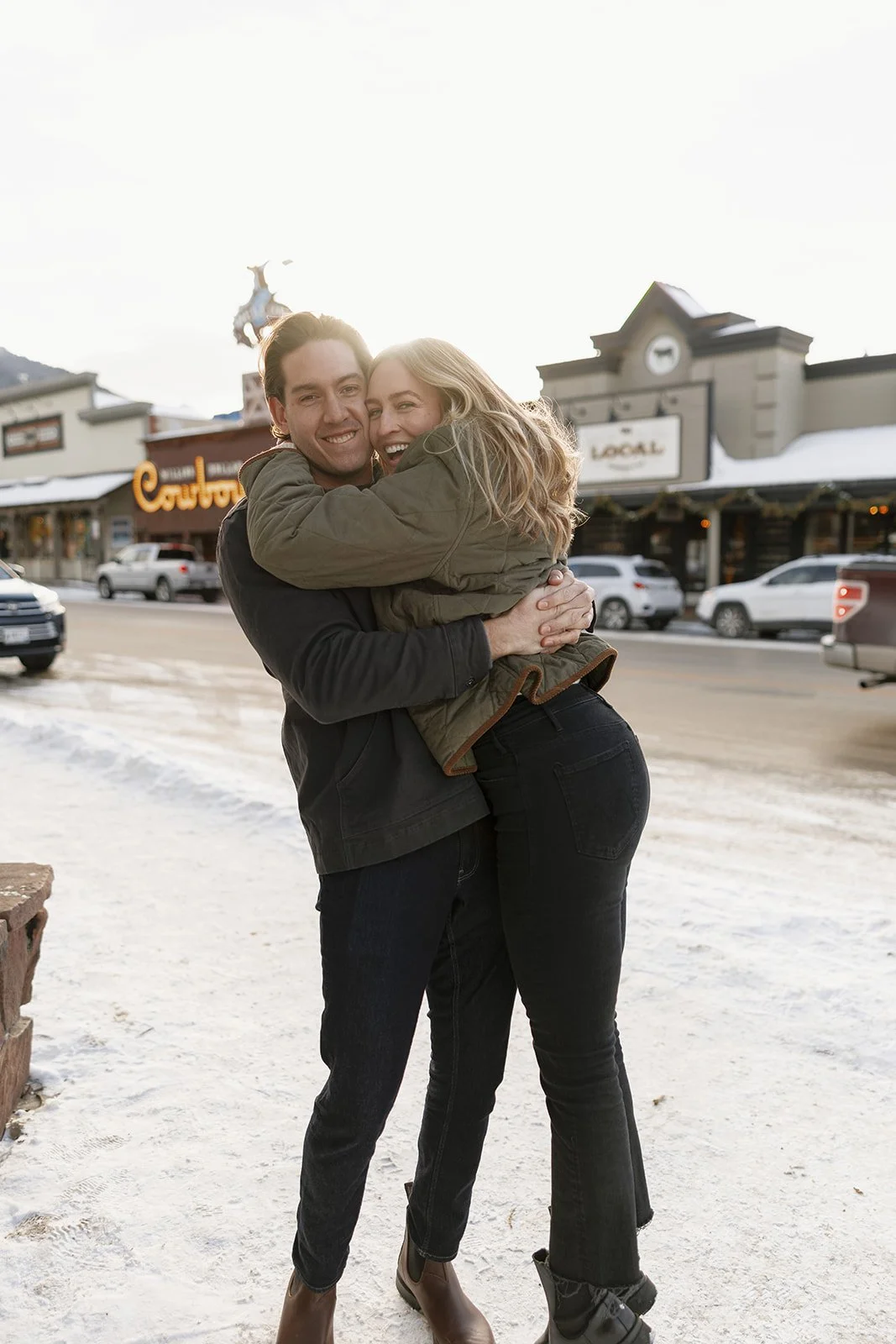 A happy couple hugging outdoors on a snowy street, smiling at the camera with shops and parked cars in the background.