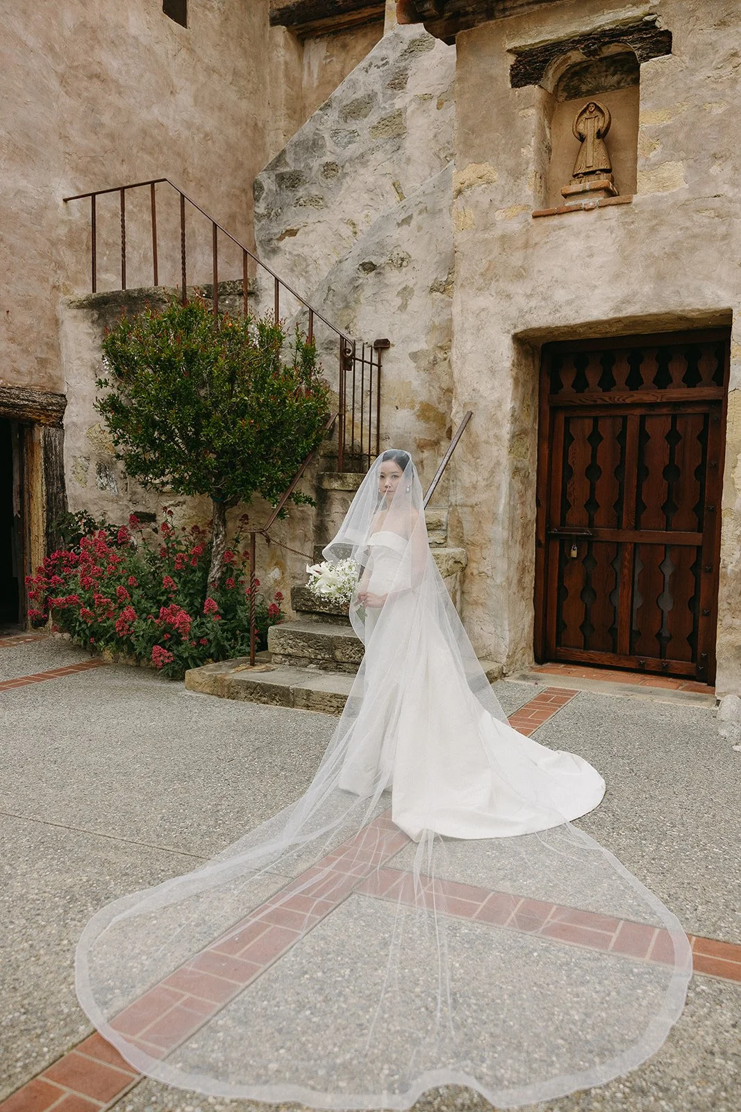 Bride in white dress and veil standing on stone steps near an old building with rustic door and wall sculpture, surrounded by red flowers.
