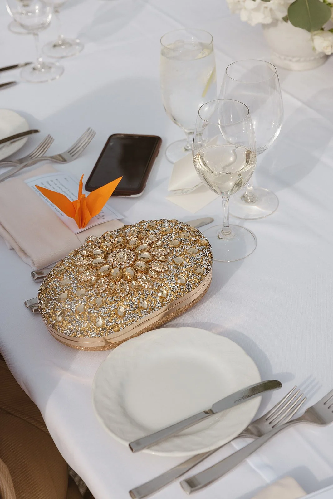 Elegant table setting with white cloth, decorative clutch, wine glasses, water glass, smartphone, orange origami crane, and floral centerpiece.