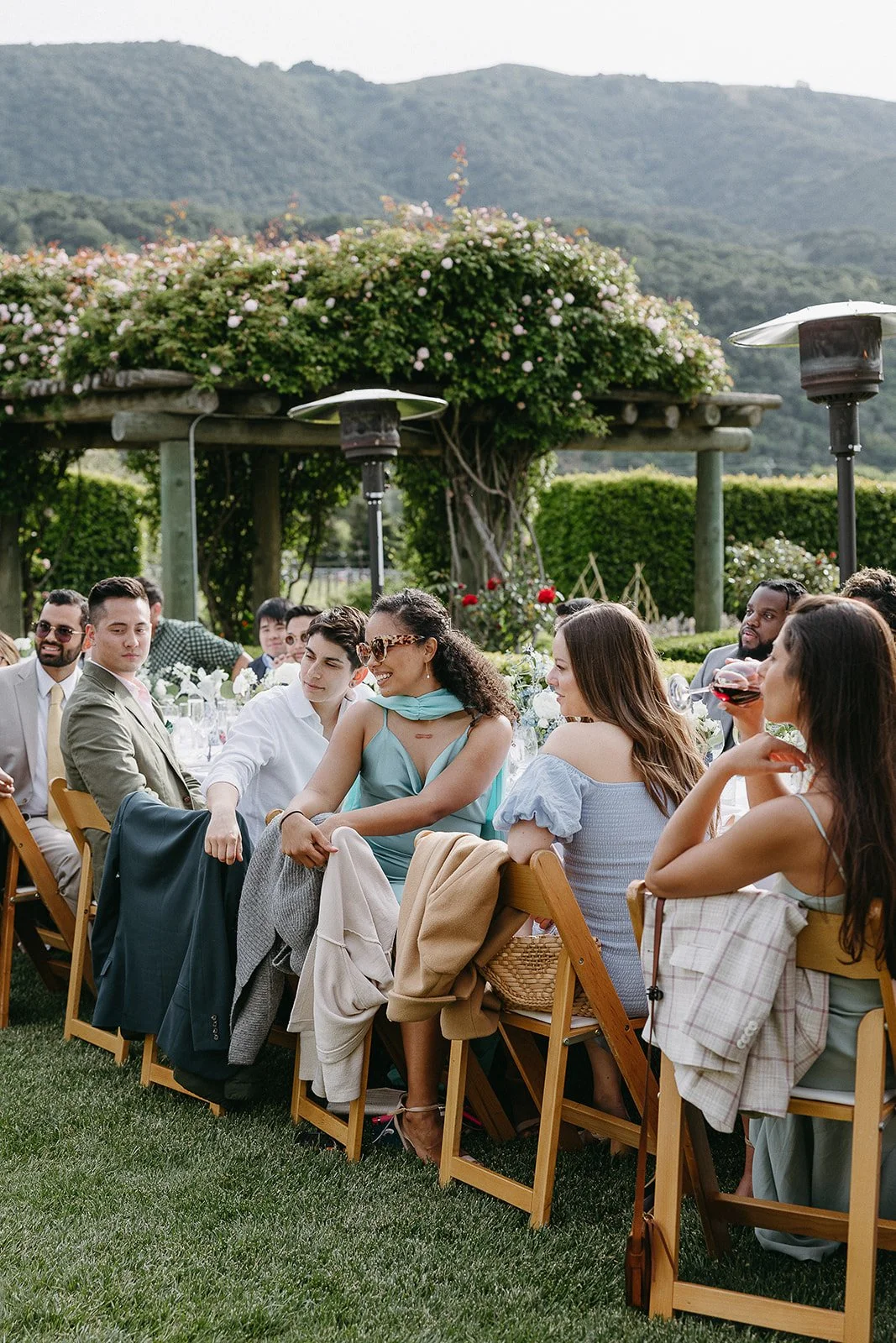 Outdoor gathering with people seated at a table, surrounded by lush greenery and hills in the background.