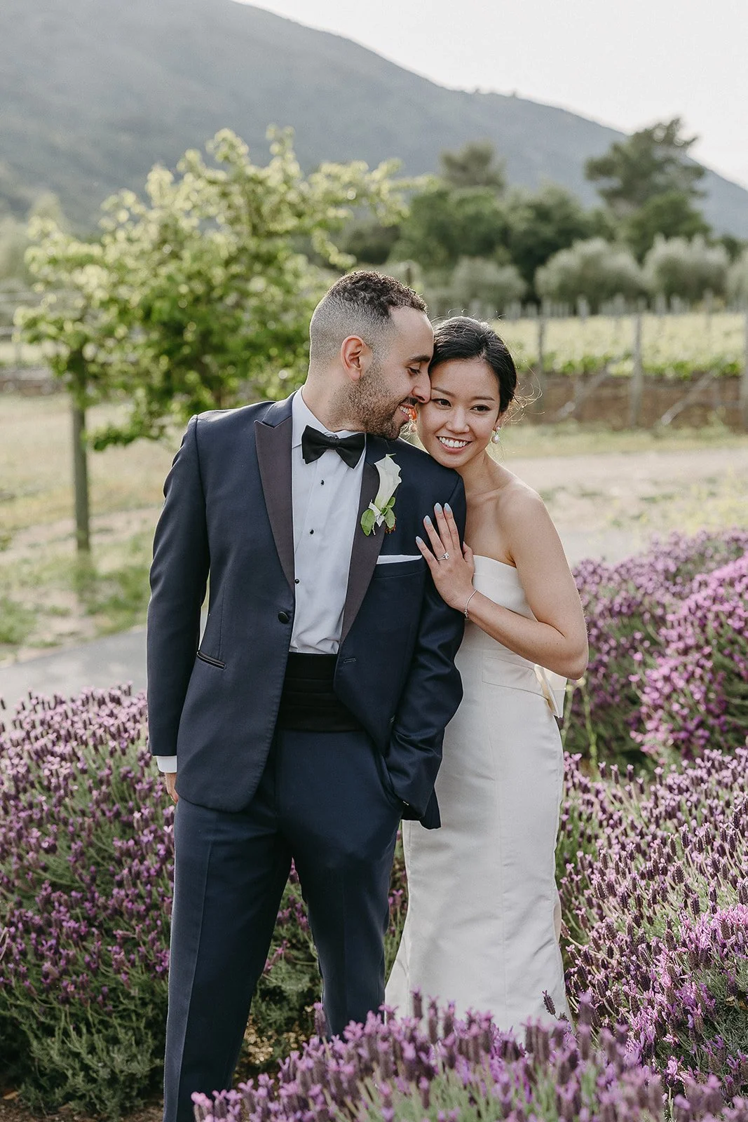 A couple dressed in formal wedding attire smiling in a garden with purple flowers, against a backdrop of hills and greenery.